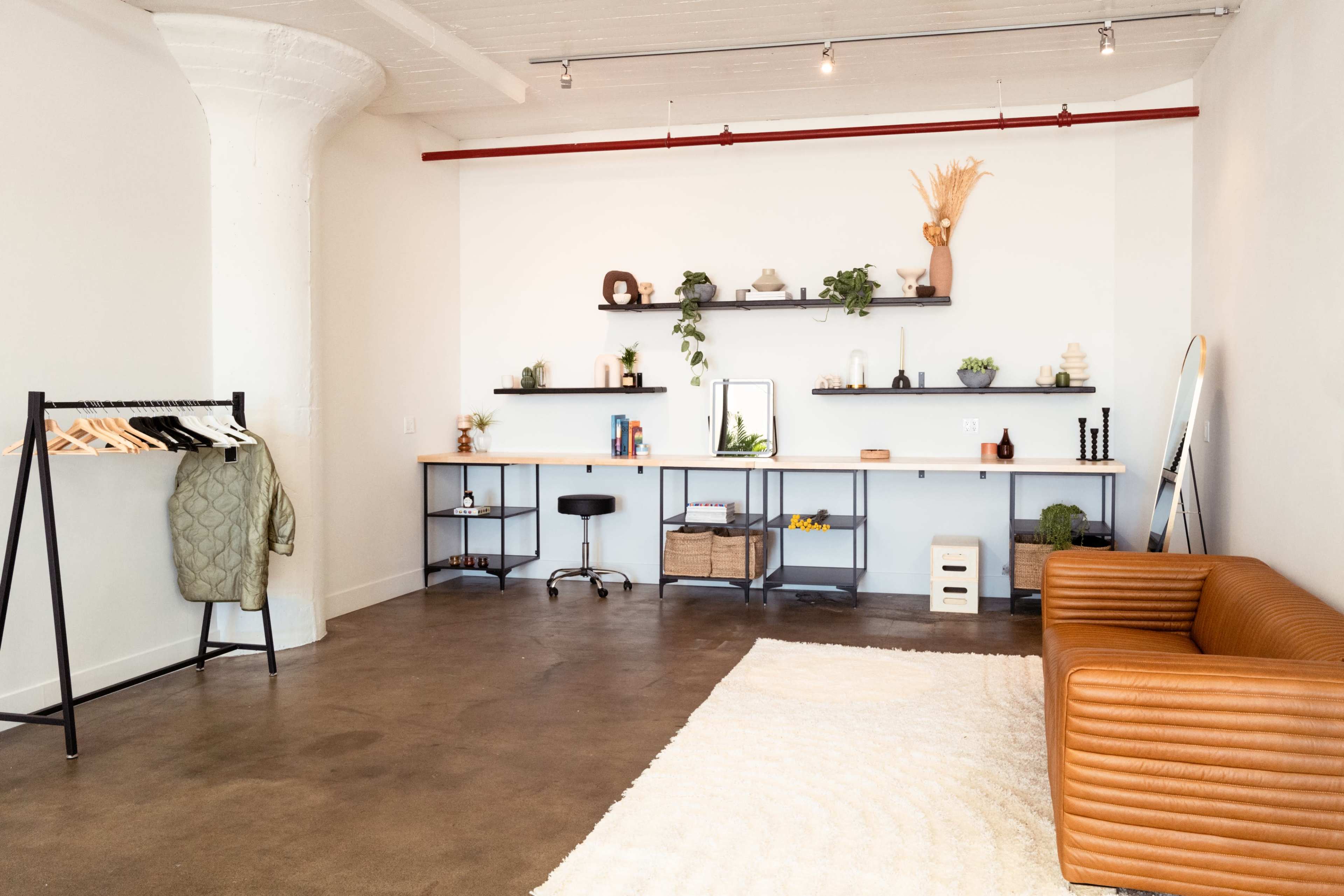 A minimalist interior space featuring a brown leather sofa, a clothing rack, a desk with shelves, and decorative plants.