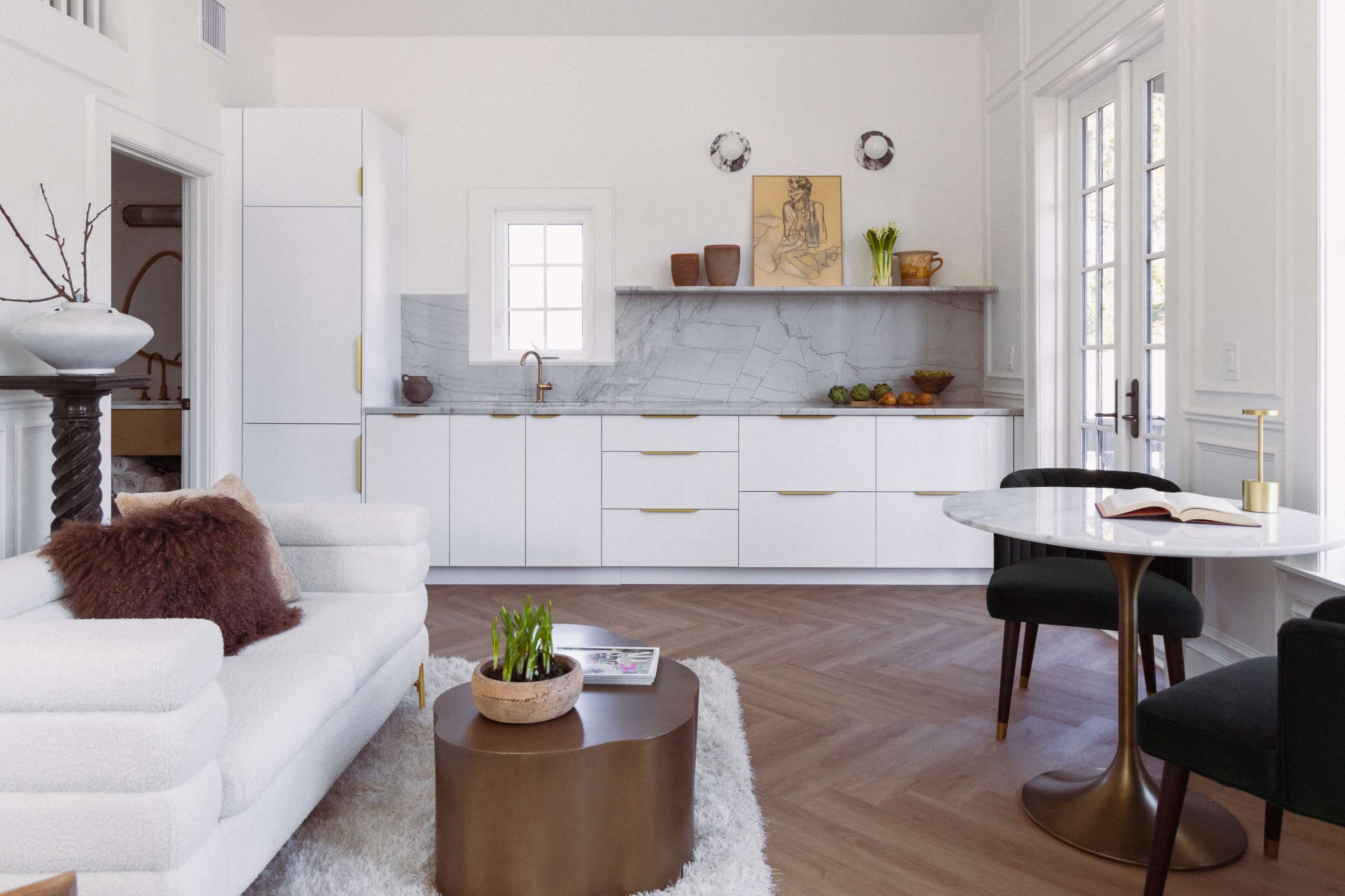 The image showcases a modern kitchen and living area with sleek white cabinetry, a marble backsplash, and a circular dining table.