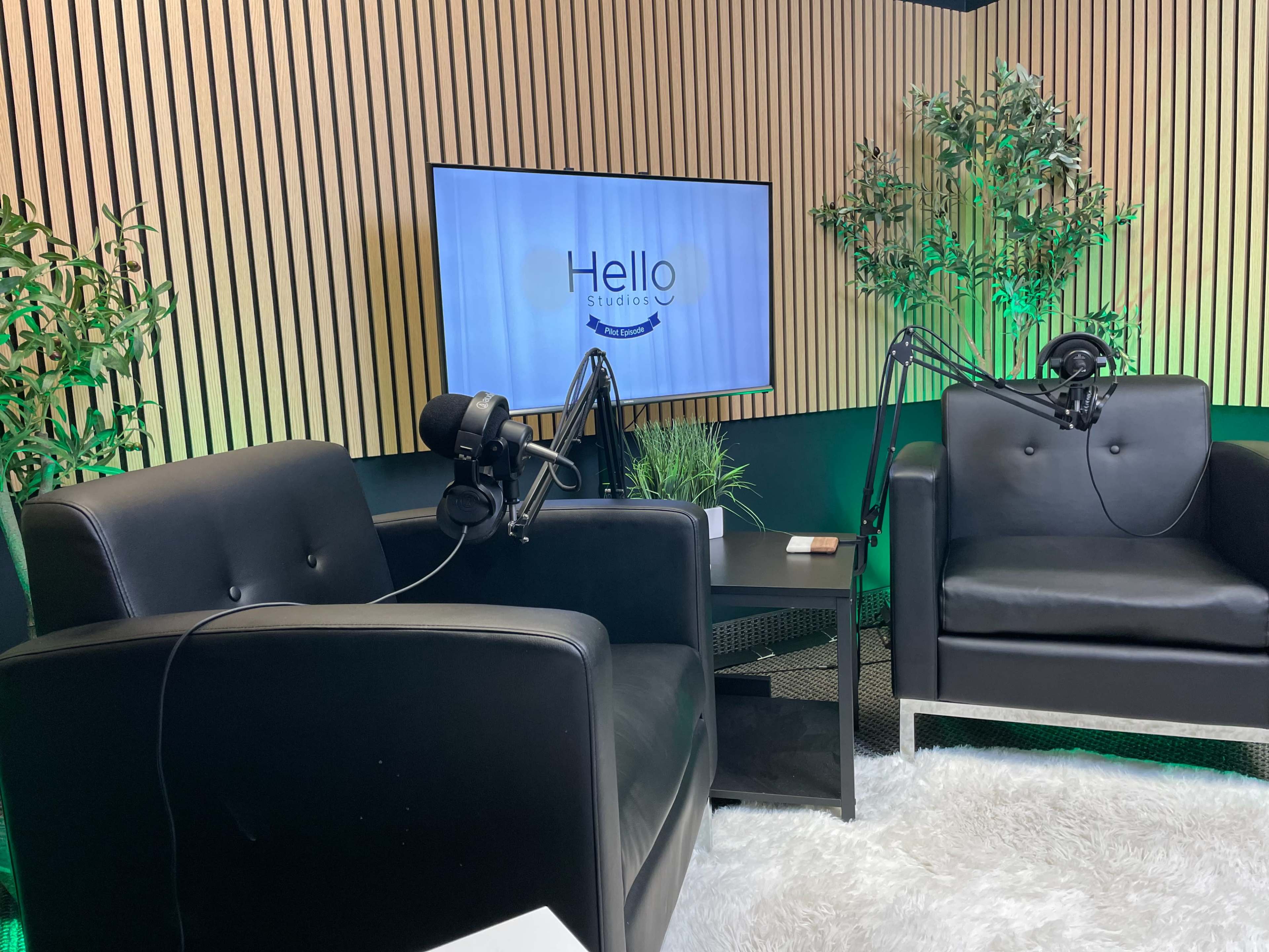 The image shows a podcast studio featuring two black chairs, a table with a microphone setup, and a television screen displaying the logo "Hello Studios."