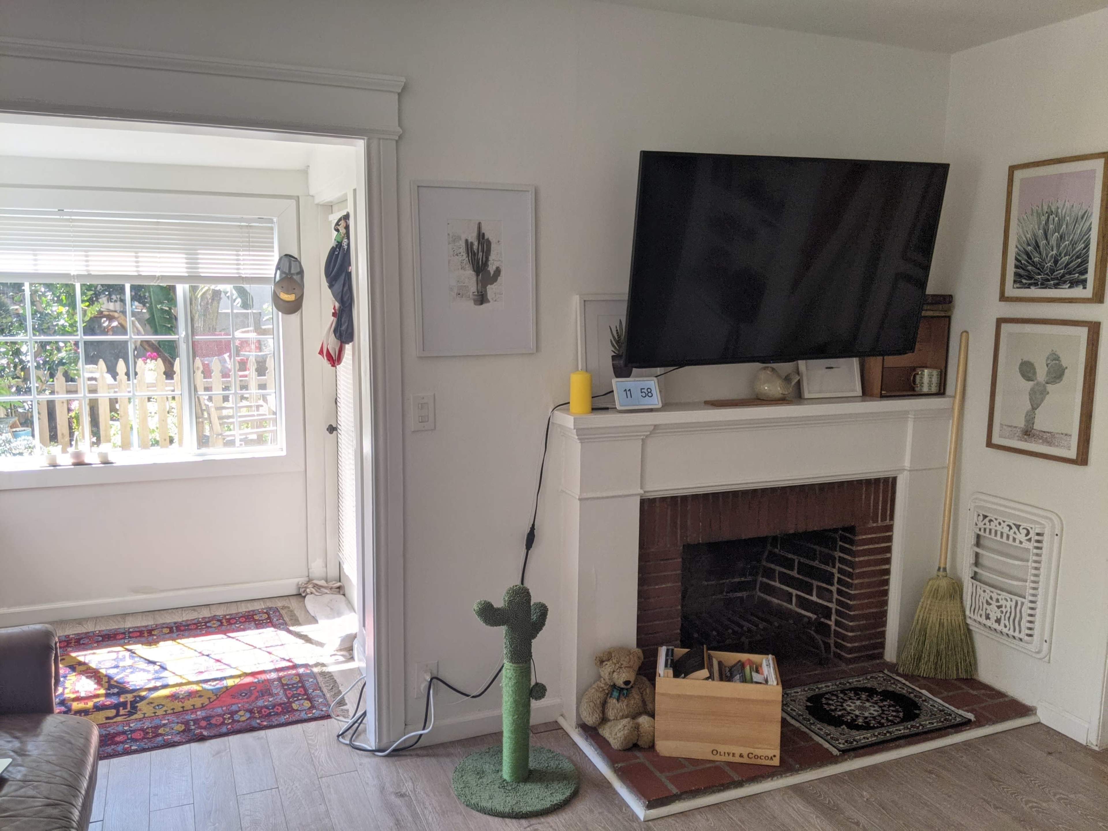 The image shows a living room featuring a large TV mounted above a fireplace, a decorative cactus, a wooden box with toys, and an adjacent sunlit space with a window.