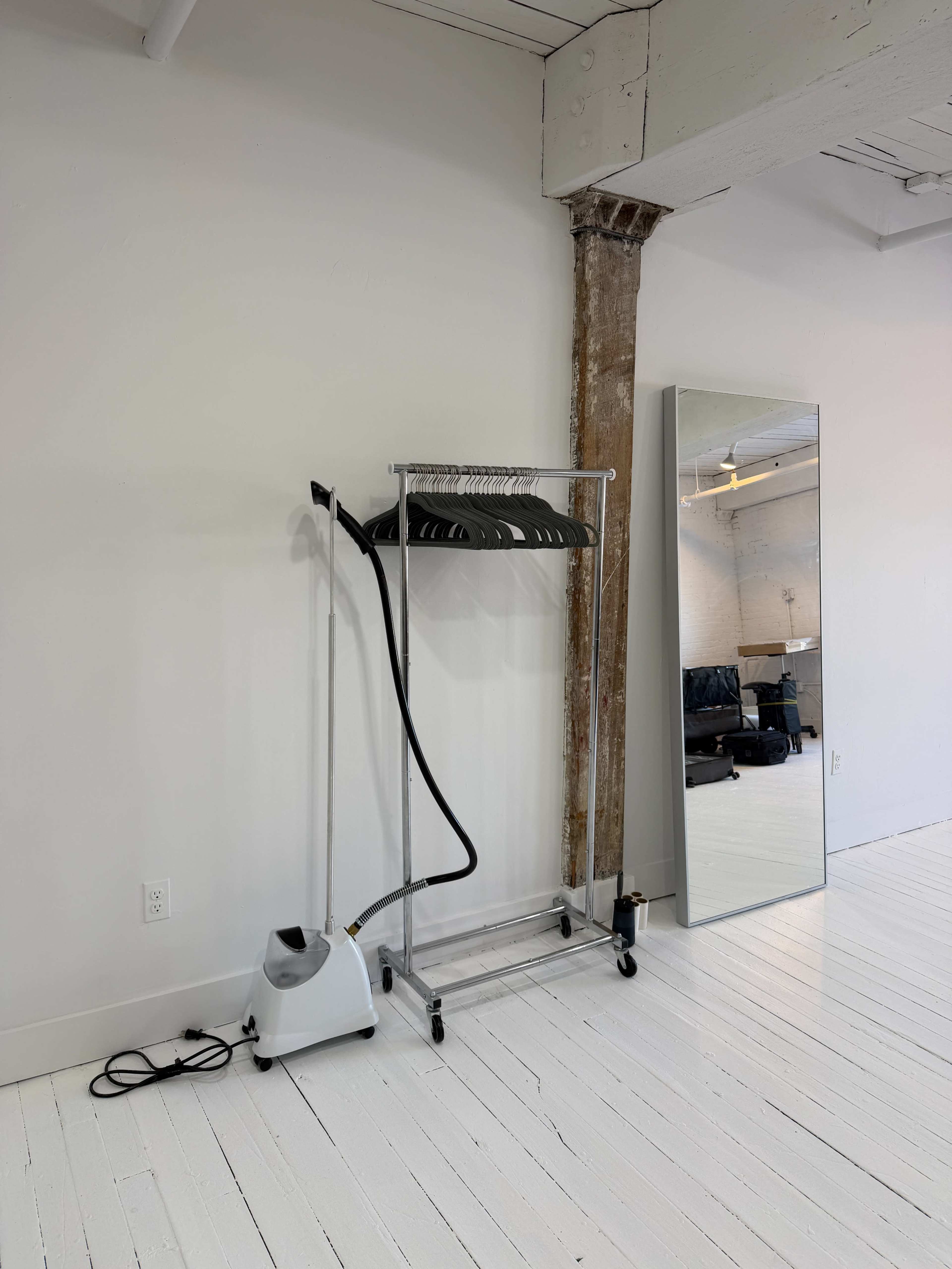 A garment rack with several hangers and a steamer stands beside a large mirror in a minimalist, well-lit room with white walls and wooden flooring.