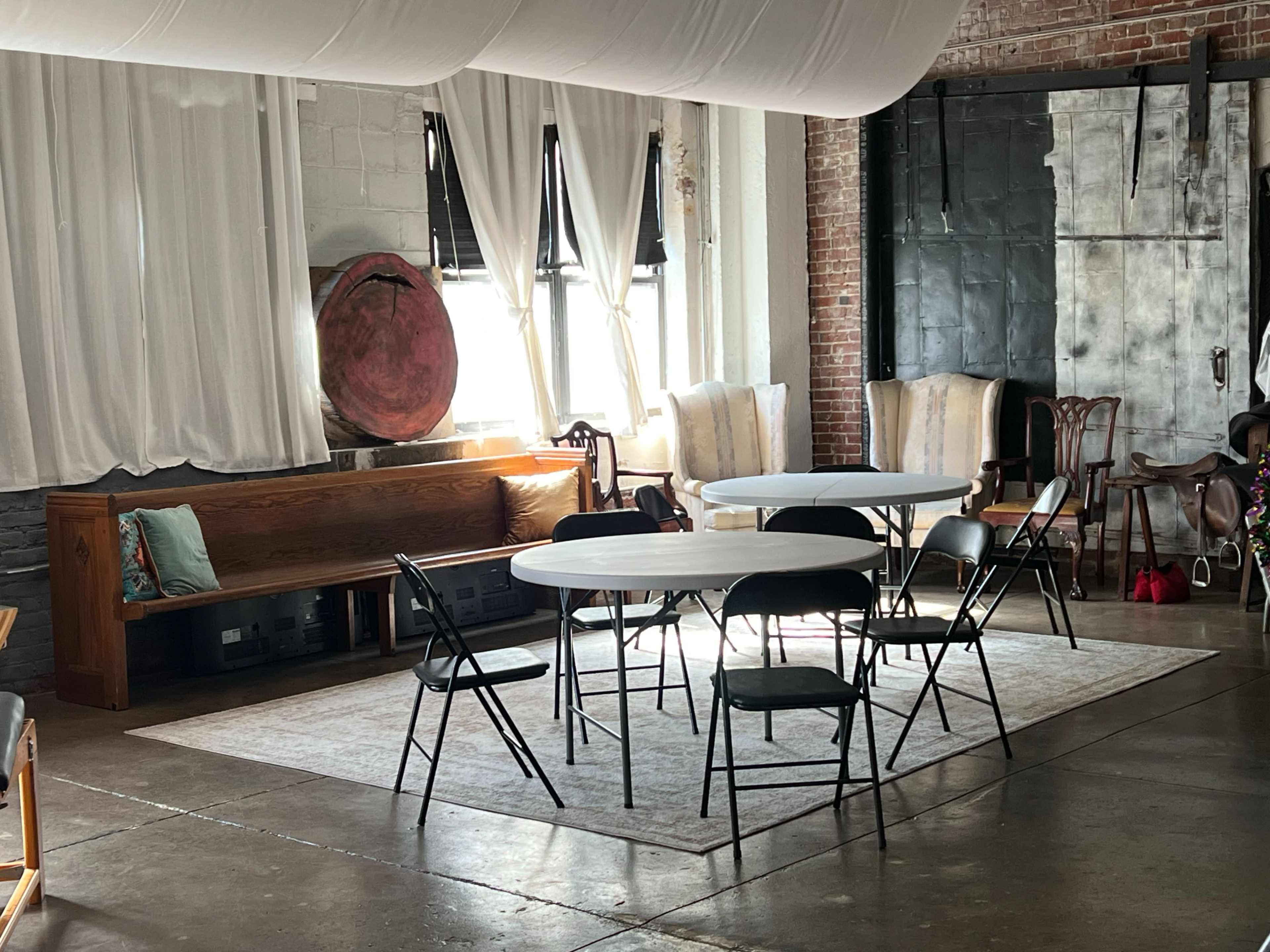 A spacious room features two round tables surrounded by black folding chairs on a light-colored rug, with large windows allowing natural light to enter.
