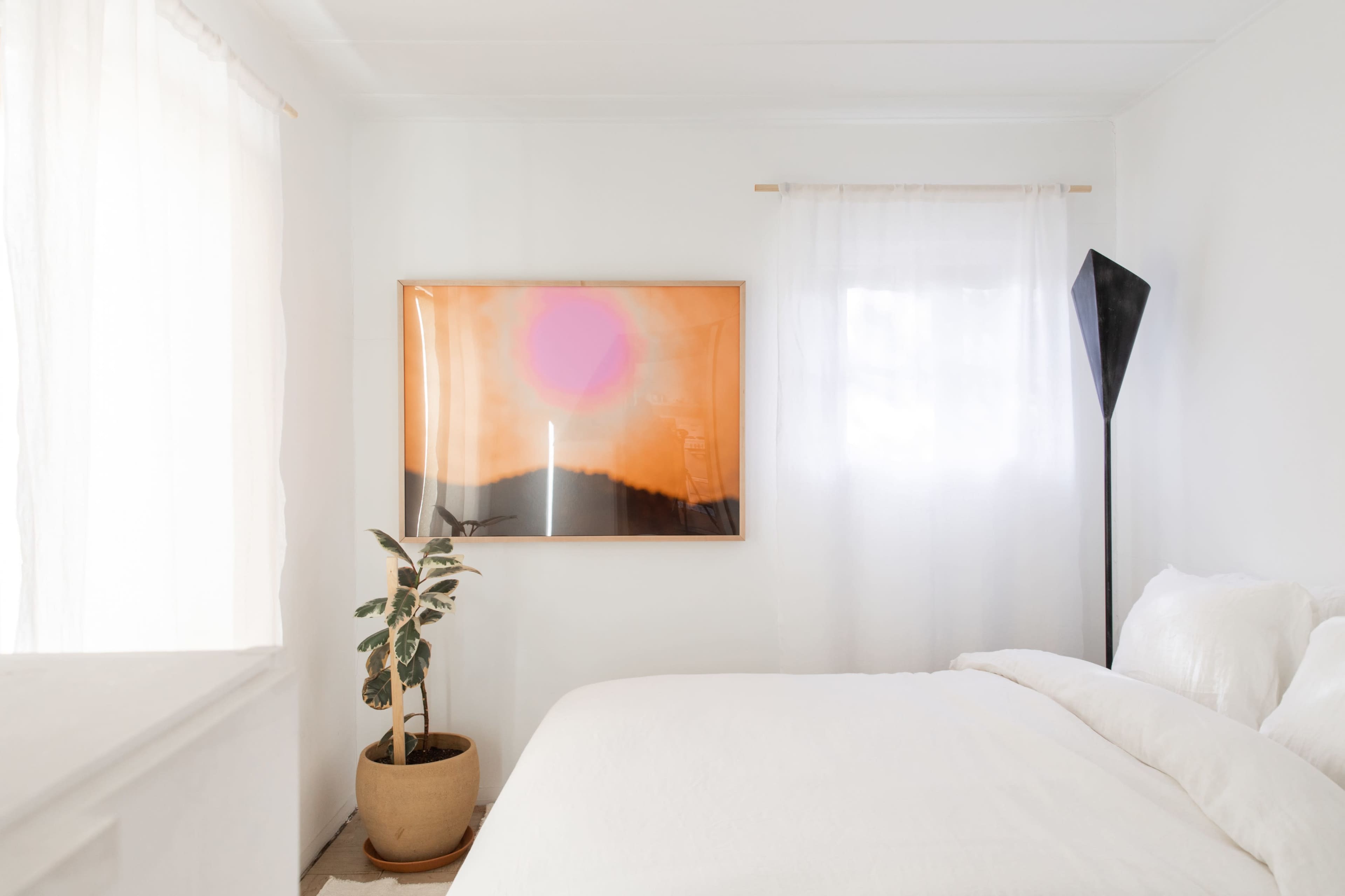 A bright bedroom features a white bed, a large orange artwork on the wall, a potted plant, and a floor lamp beside a window.