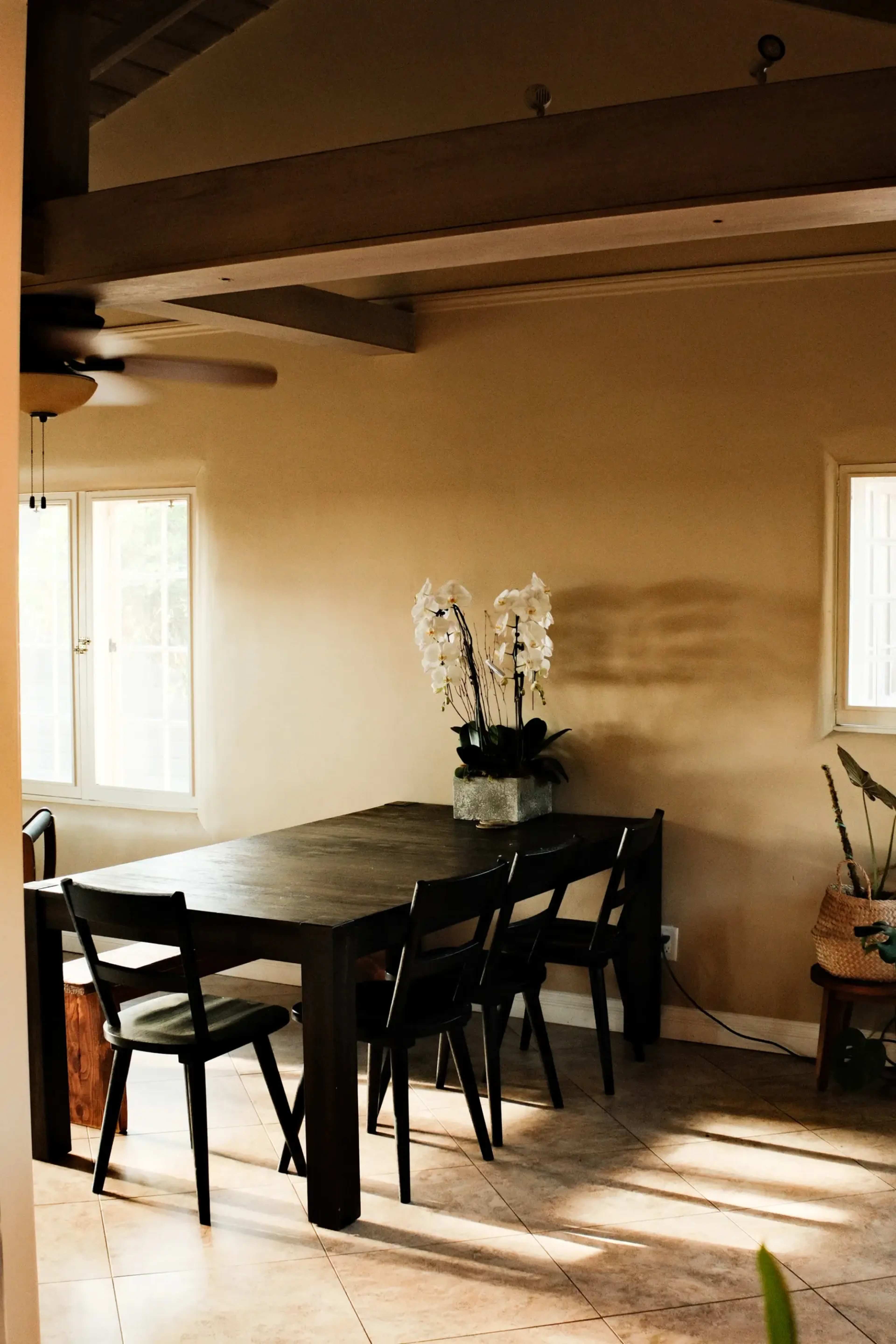 A dining area features a wooden table surrounded by black chairs, with a potted orchid arrangement and large windows allowing natural light to enter.