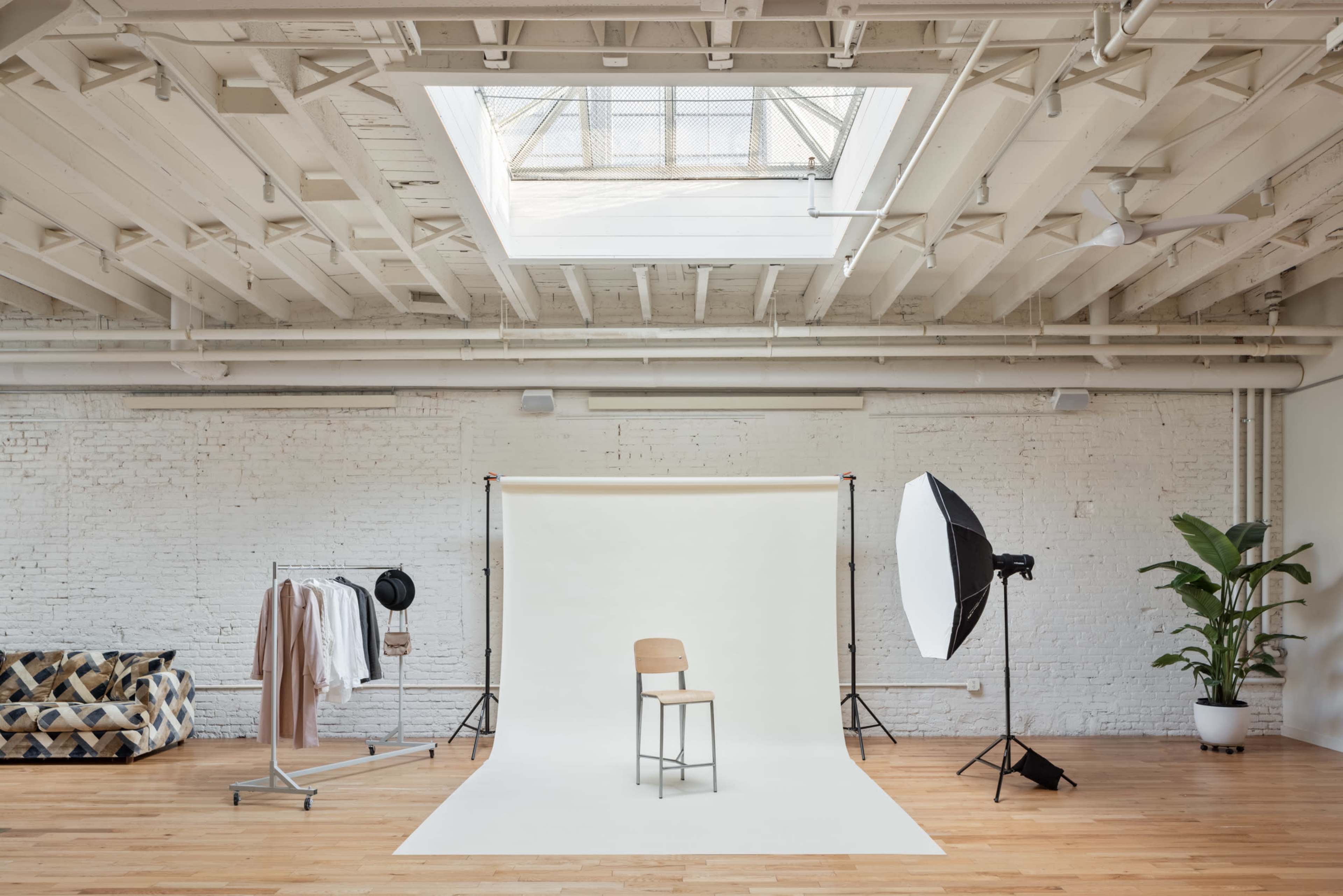 A photography studio featuring a wooden chair on a white backdrop, surrounded by clothing on a rack, a large light source, and a potted plant.