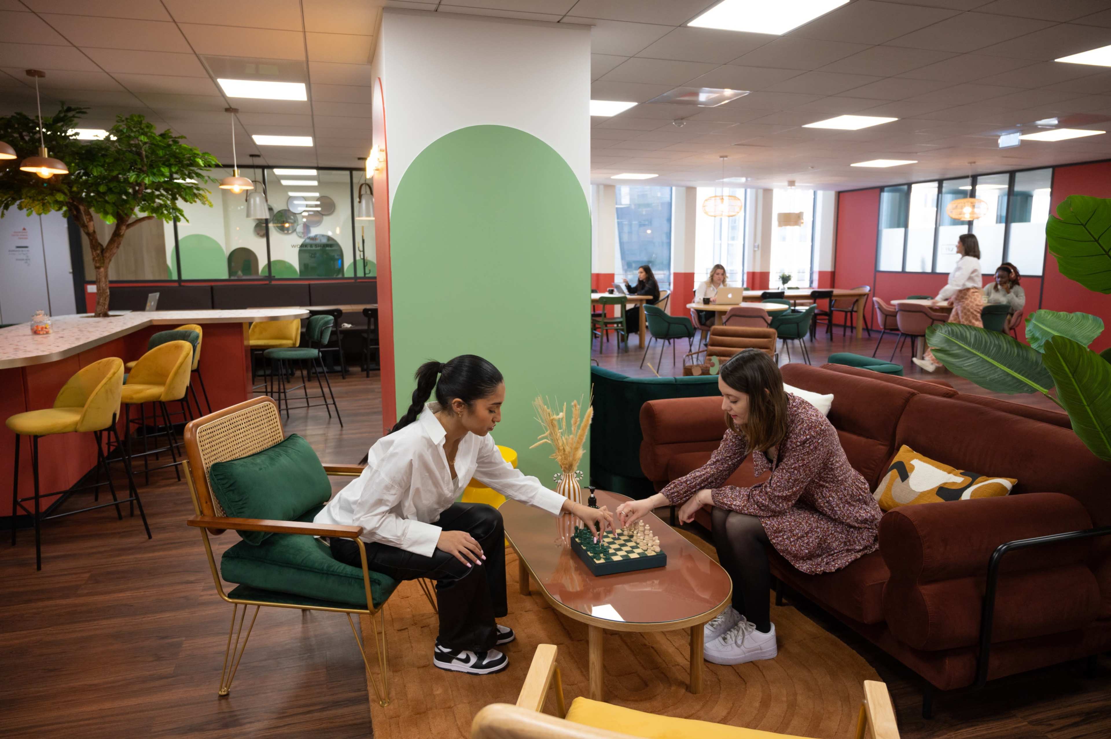 Two women play chess at a coffee table in a modern, open-plan office lounge with colorful seating and greenery.
