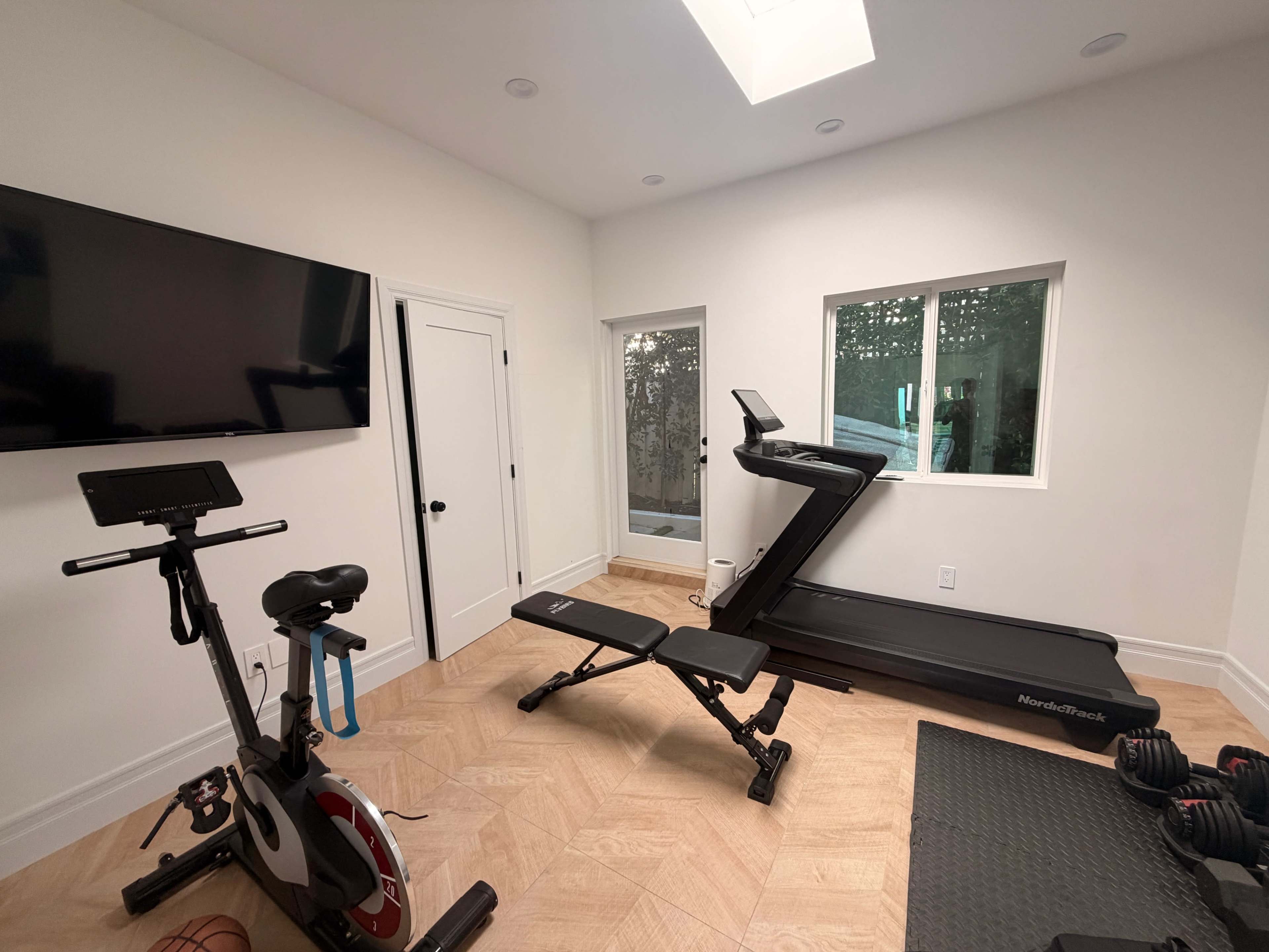 The image shows a modern home gym featuring a treadmill, a stationary bike, free weights, and a large television mounted on the wall.