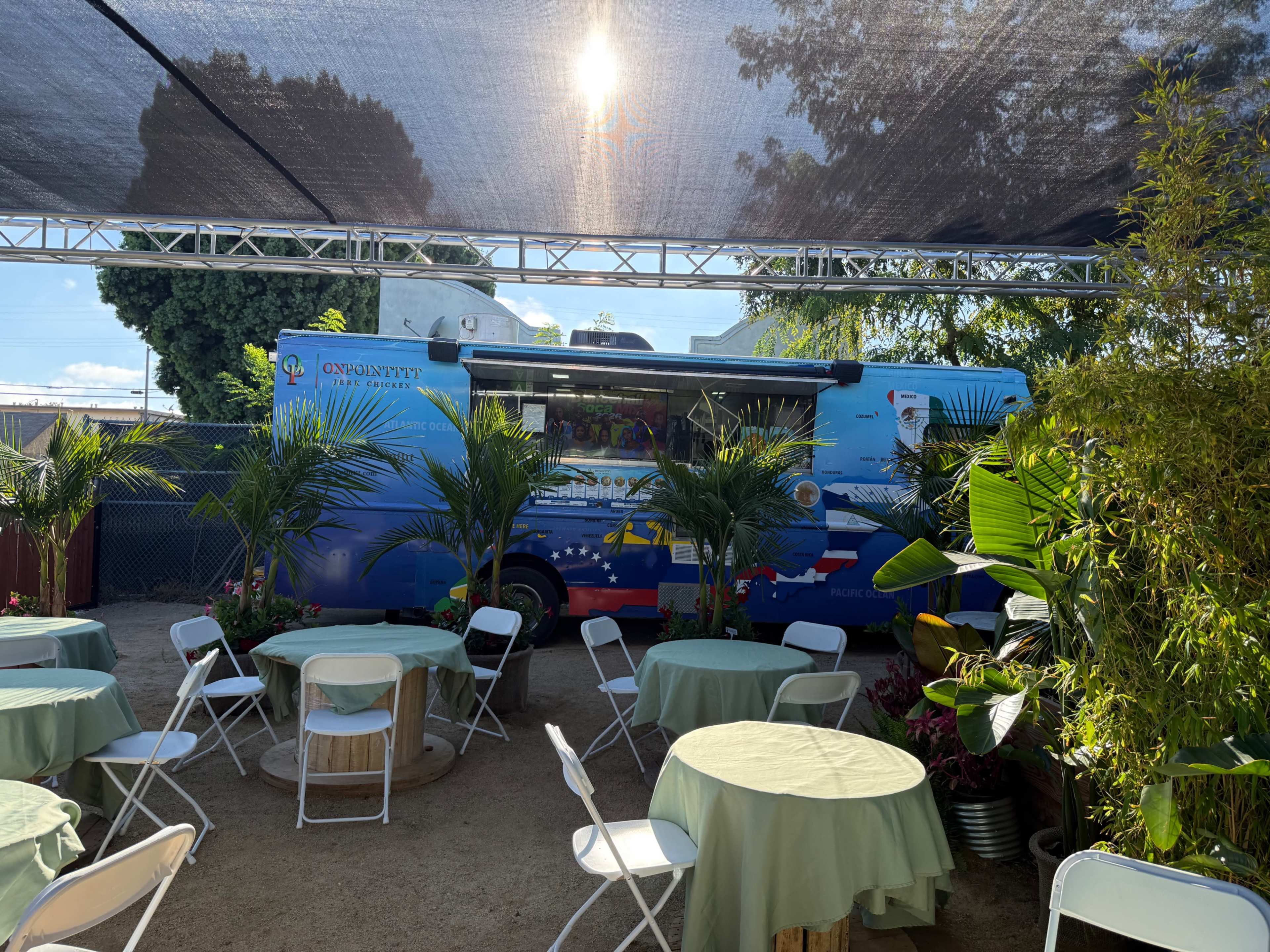 A food truck is parked under a shaded area surrounded by tables and decorative plants.