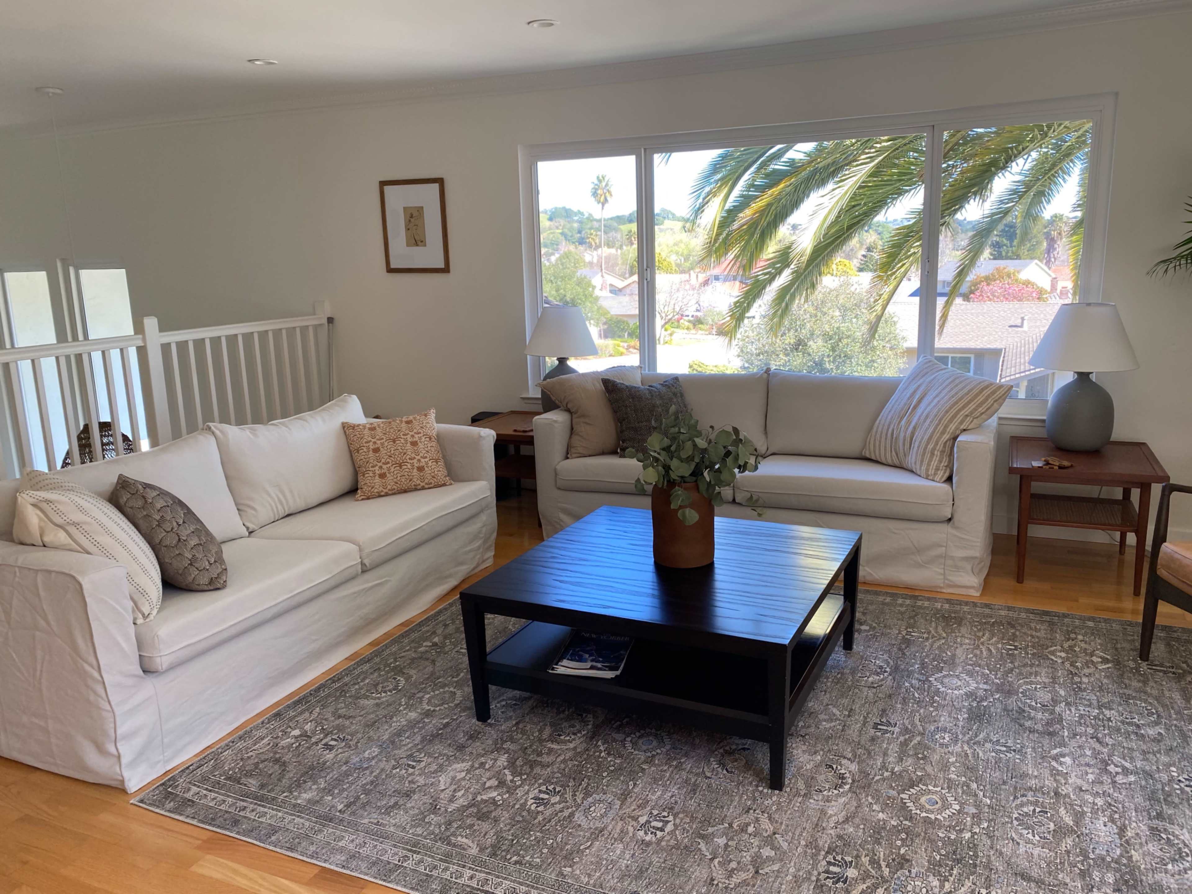 A living room features two white sofas, a black coffee table, and large windows with a view of palm trees.