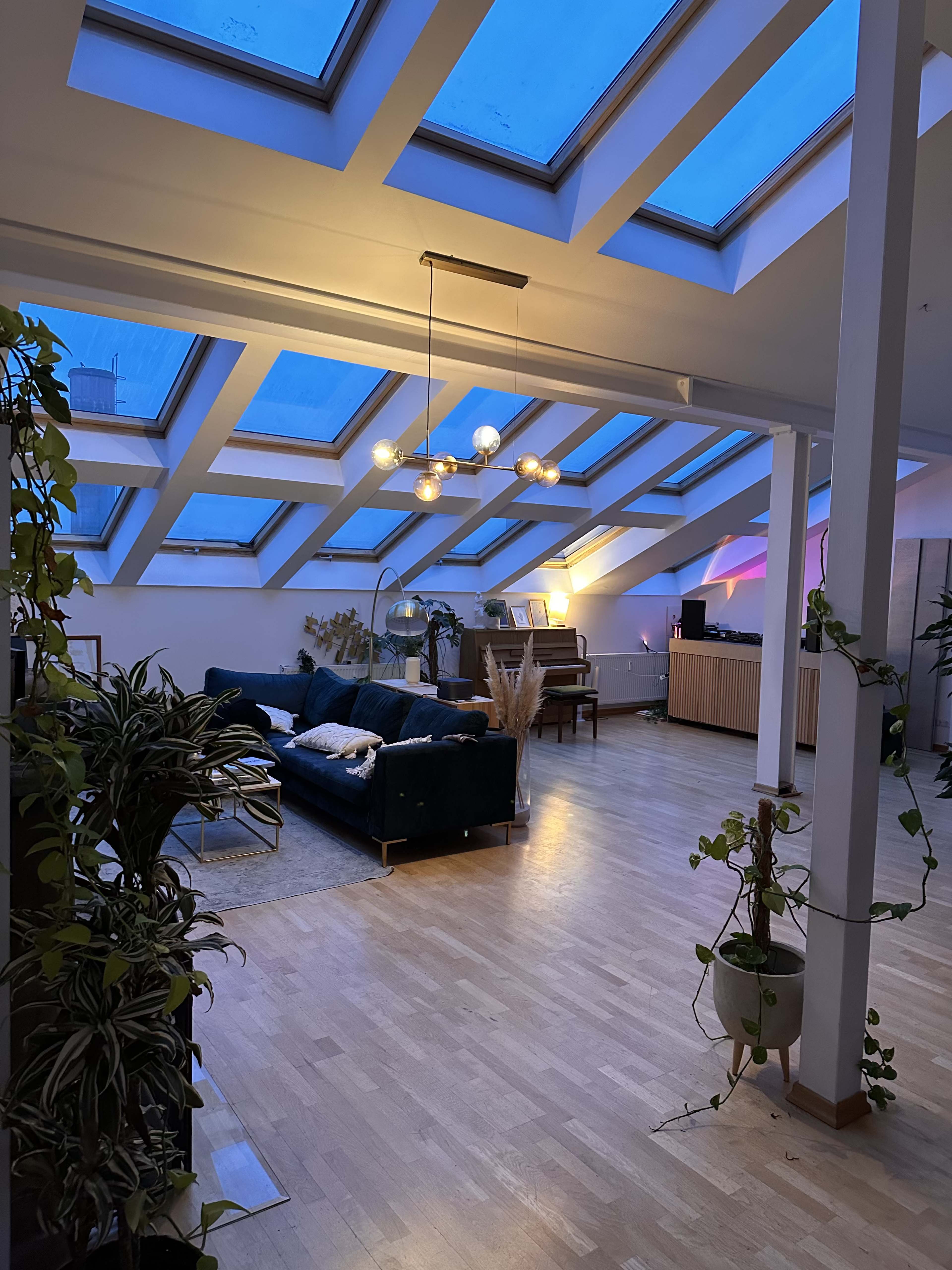 The image shows a spacious room with large skylights and wooden flooring, featuring a dark sofa, plants, and a music setup in a modern interior.