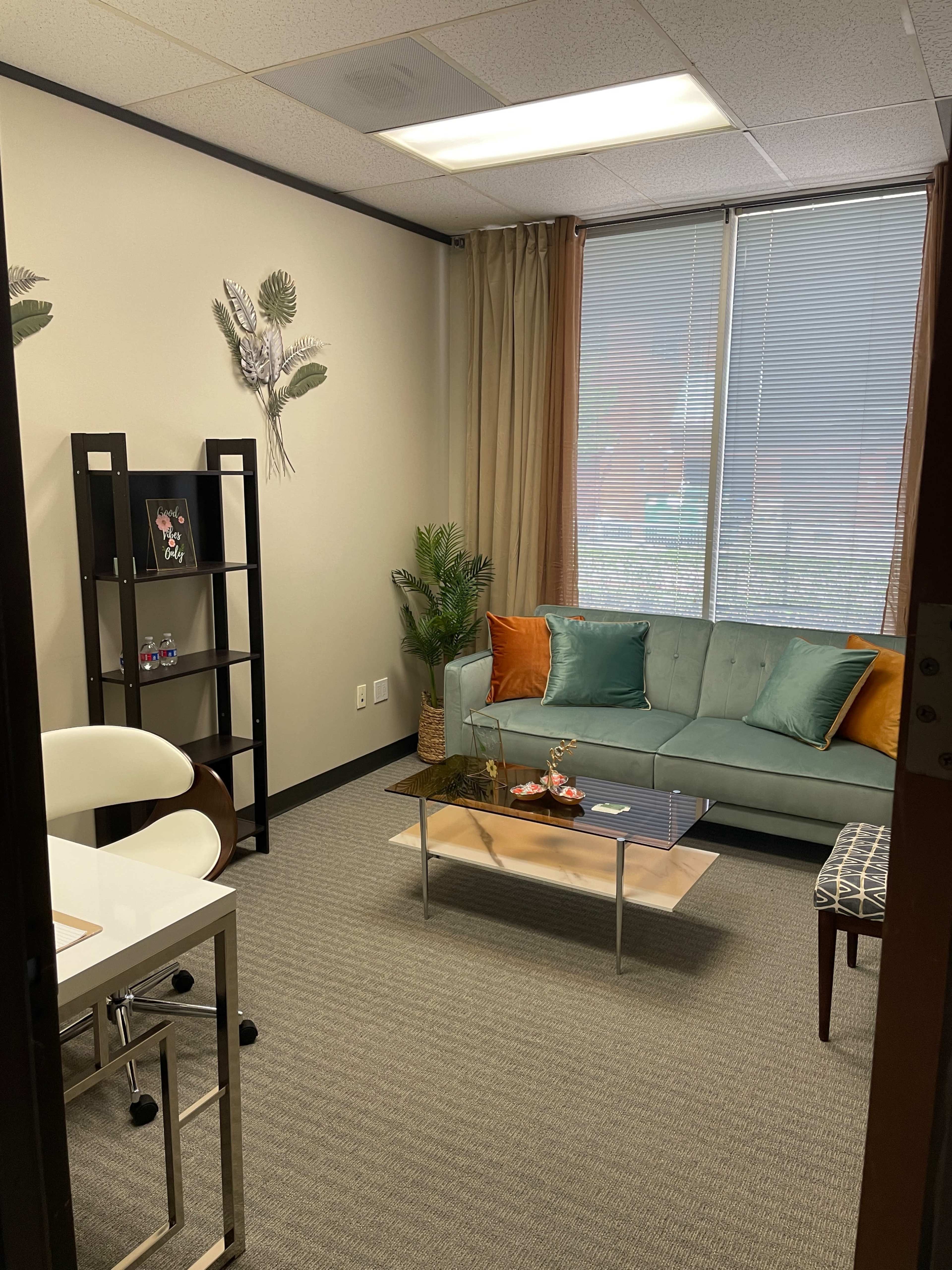 The image shows a small office space with a teal sofa, a coffee table, and a black shelving unit, all arranged within a well-lit room featuring a window.