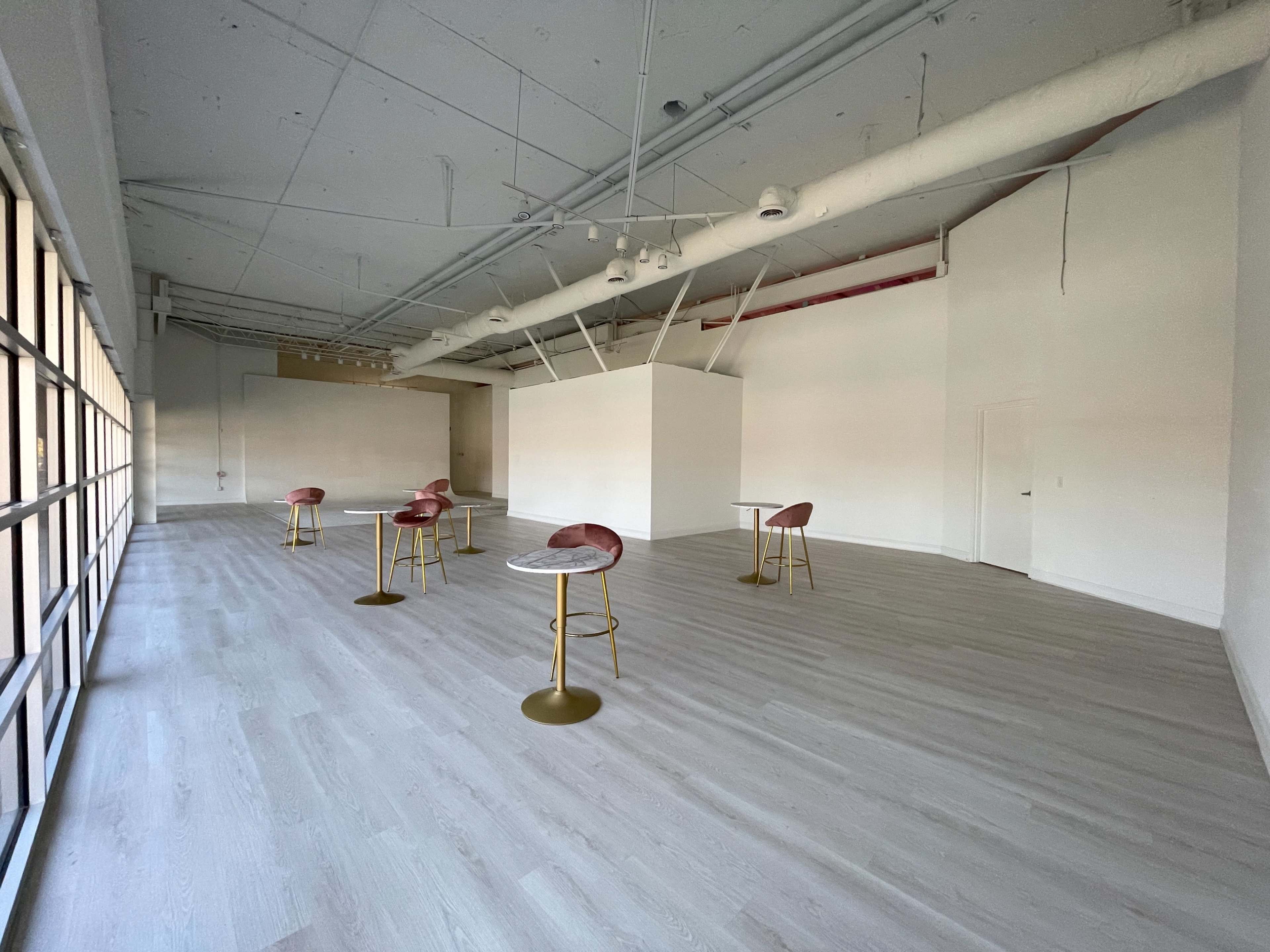 An empty modern room features several tall small tables with pink seating arranged on a light-colored wooden floor.