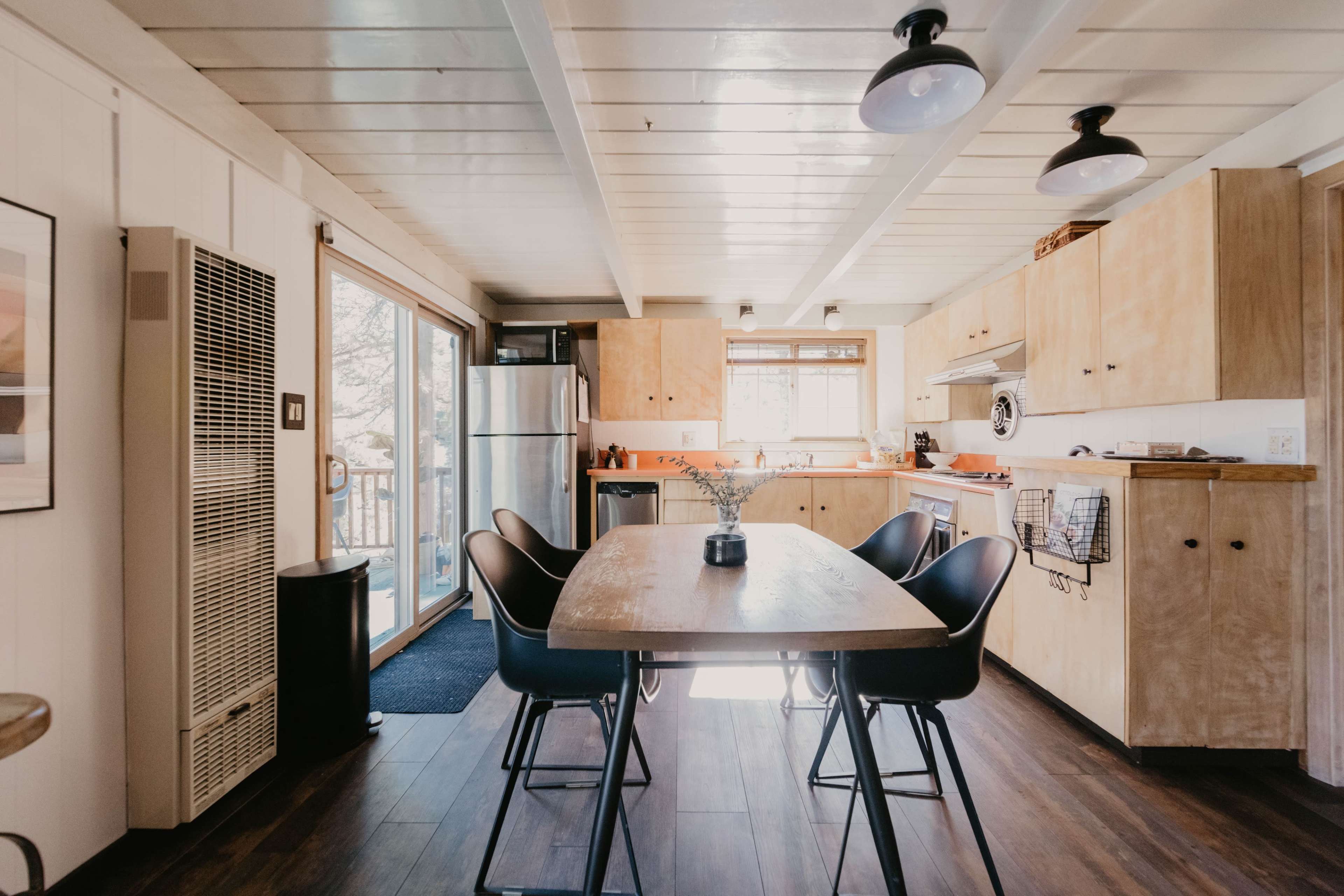 A modern kitchen features a wooden table surrounded by black chairs, with stainless steel appliances and sliding doors leading to an outdoor area.