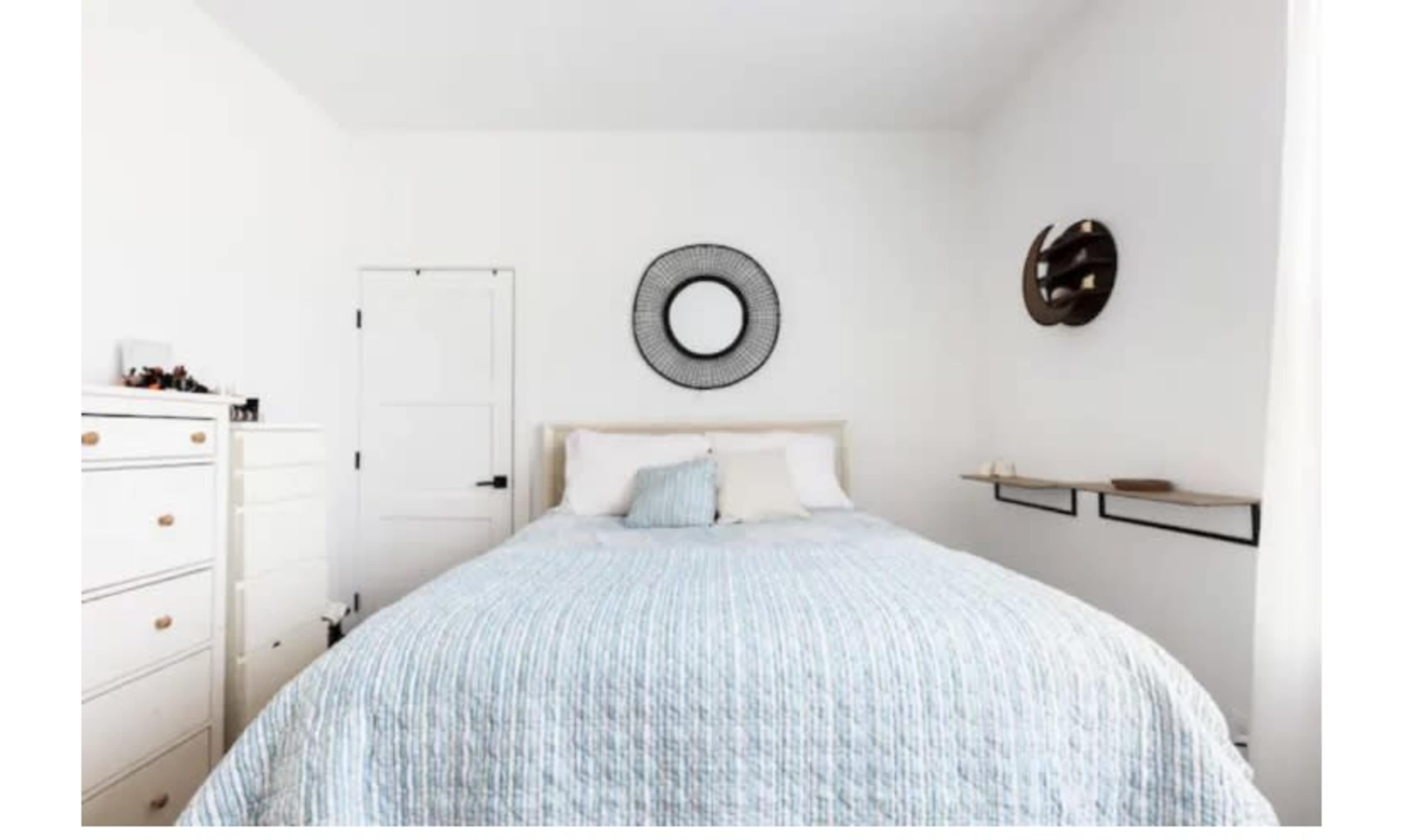 A minimalist bedroom with a bed covered in a light blue quilt, a round mirror on the wall, and a small shelf above a dresser.