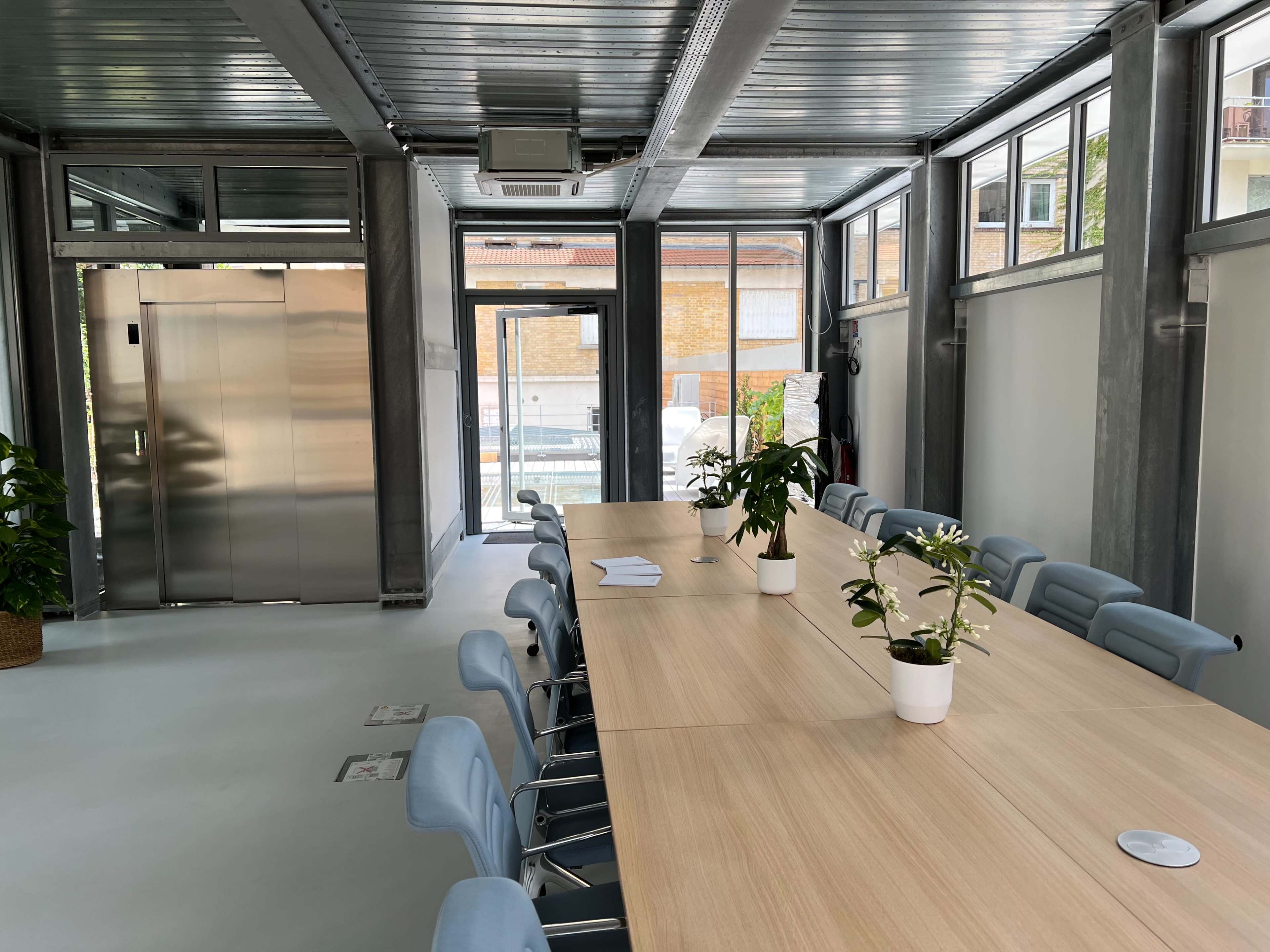A large conference room features a long wooden table surrounded by light blue chairs and plants, with glass walls and an elevator visible in the background.