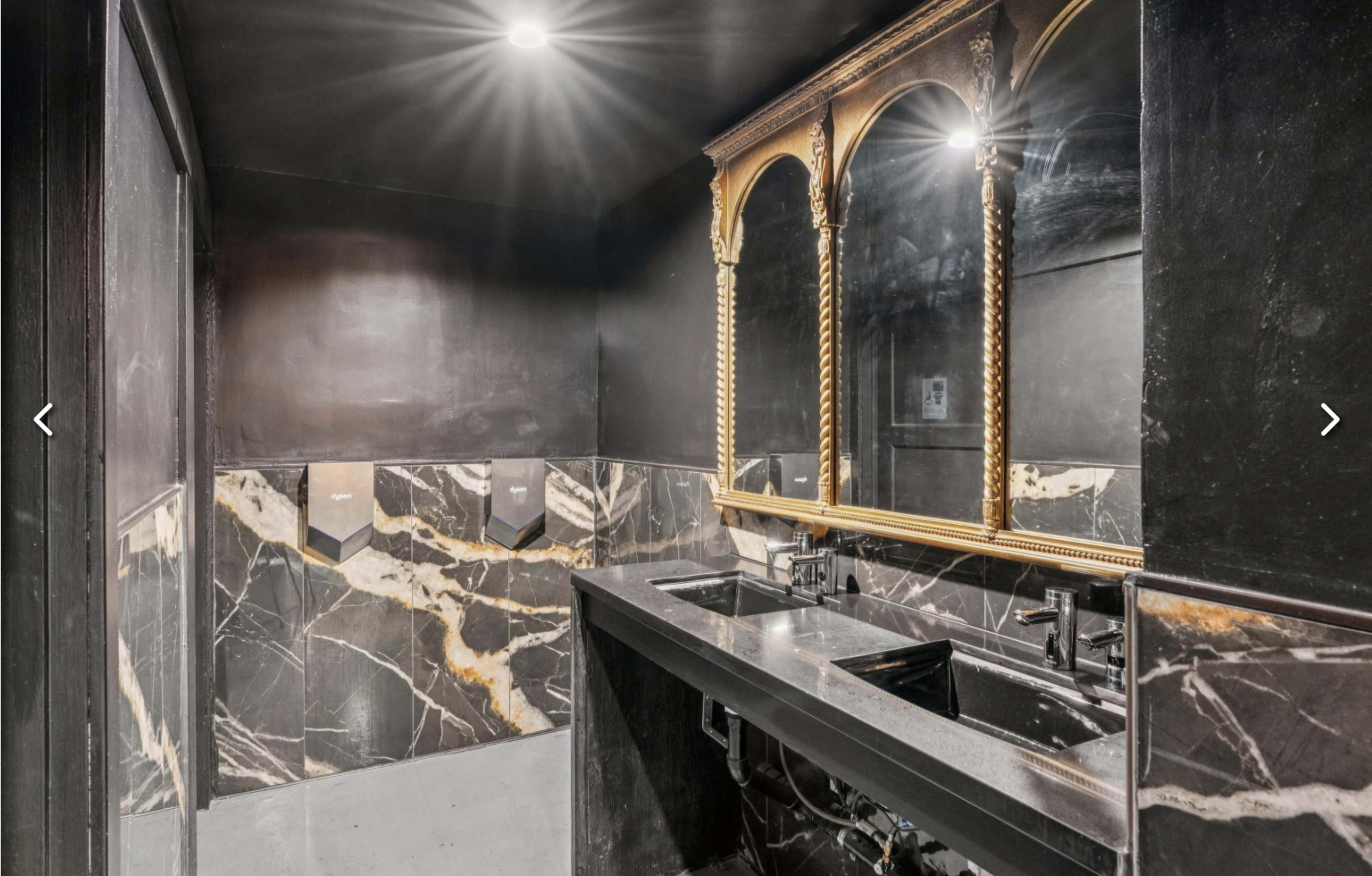 The image shows a modern bathroom with dark marble walls, a black countertop featuring dual sinks, and a decorative mirror mounted above.