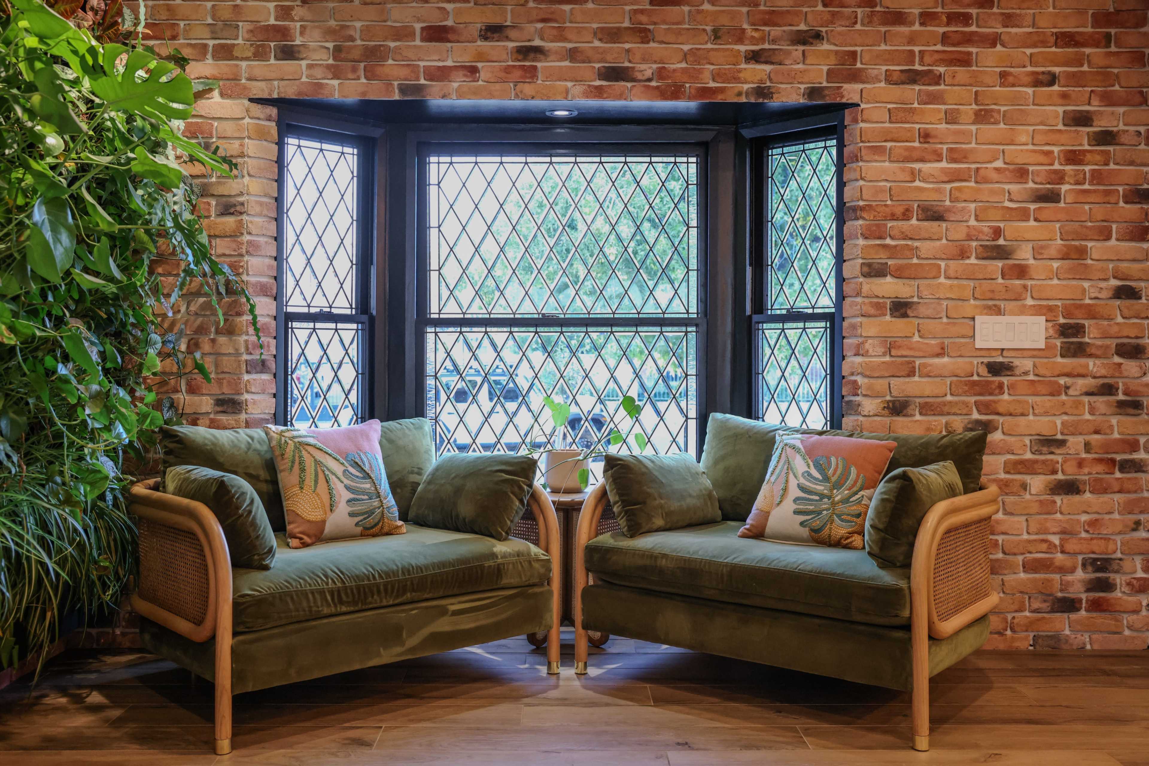 Two green sofas with decorative pillows sit in a cozy nook beside large windows, surrounded by a brick wall and indoor plants.