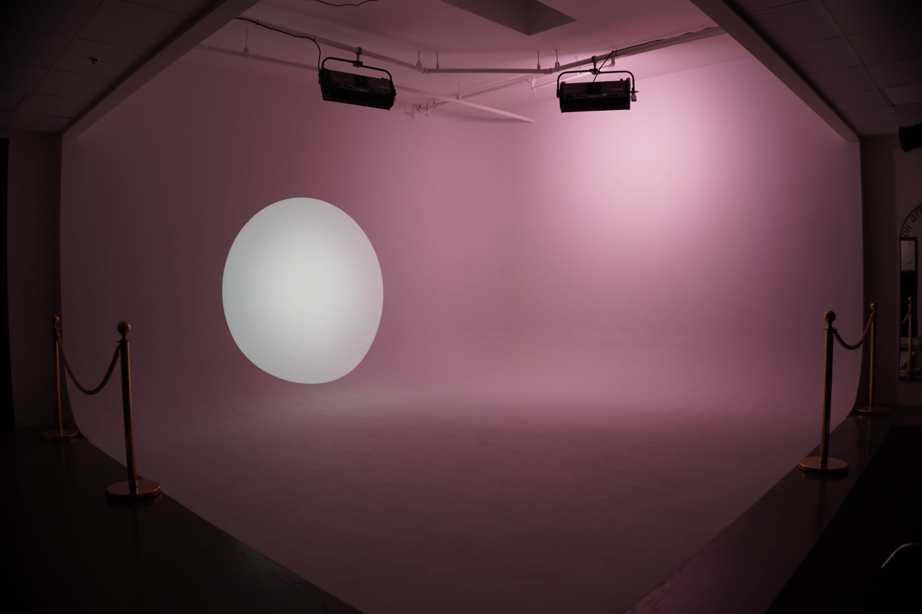 The image shows a minimalist studio space with a soft pink backdrop, featuring a large white circular light projection on one side.