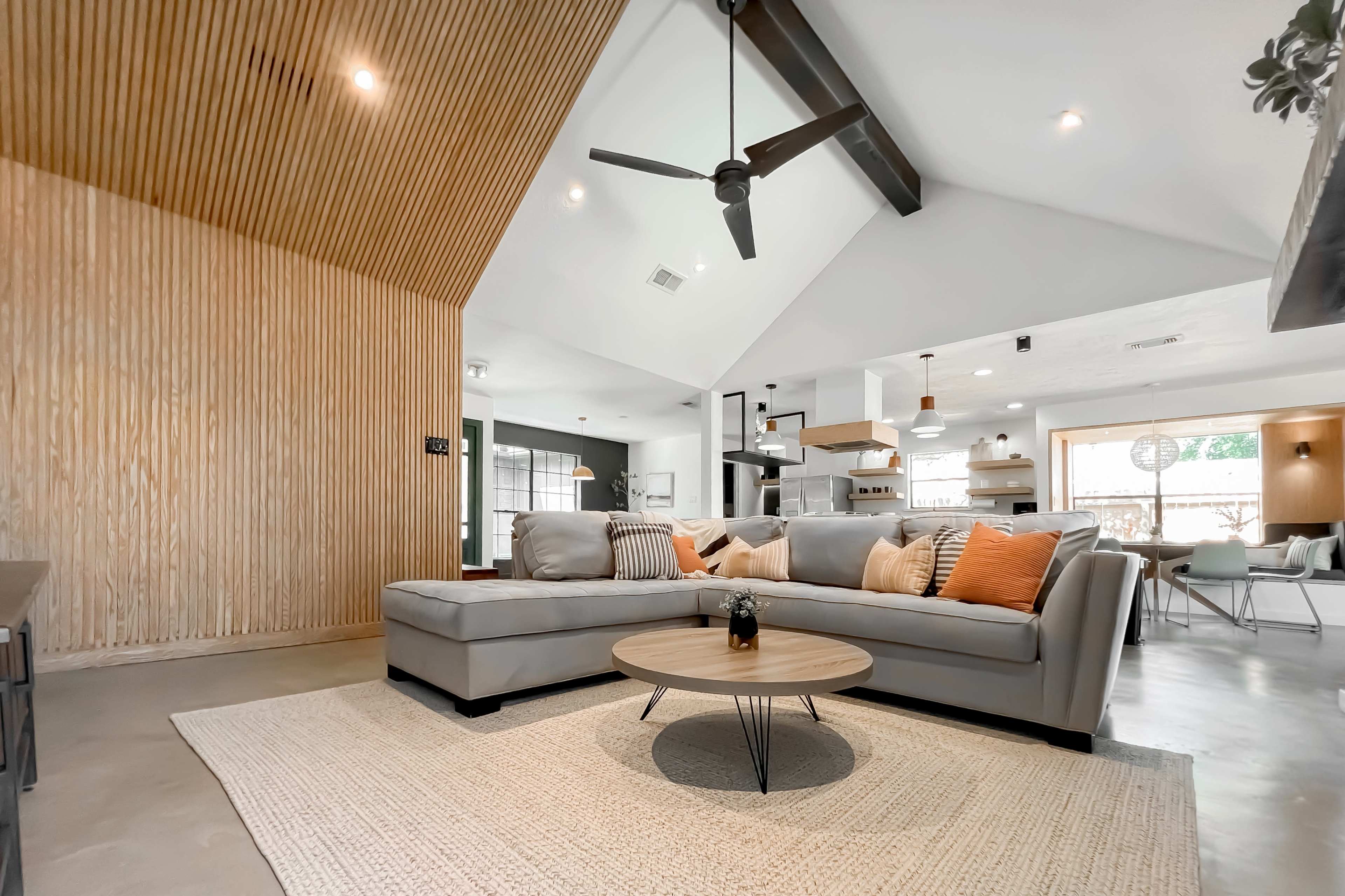 A modern living room with a light gray sectional sofa, a circular coffee table, and a textured wooden accent wall, all under a high ceiling with exposed beams.