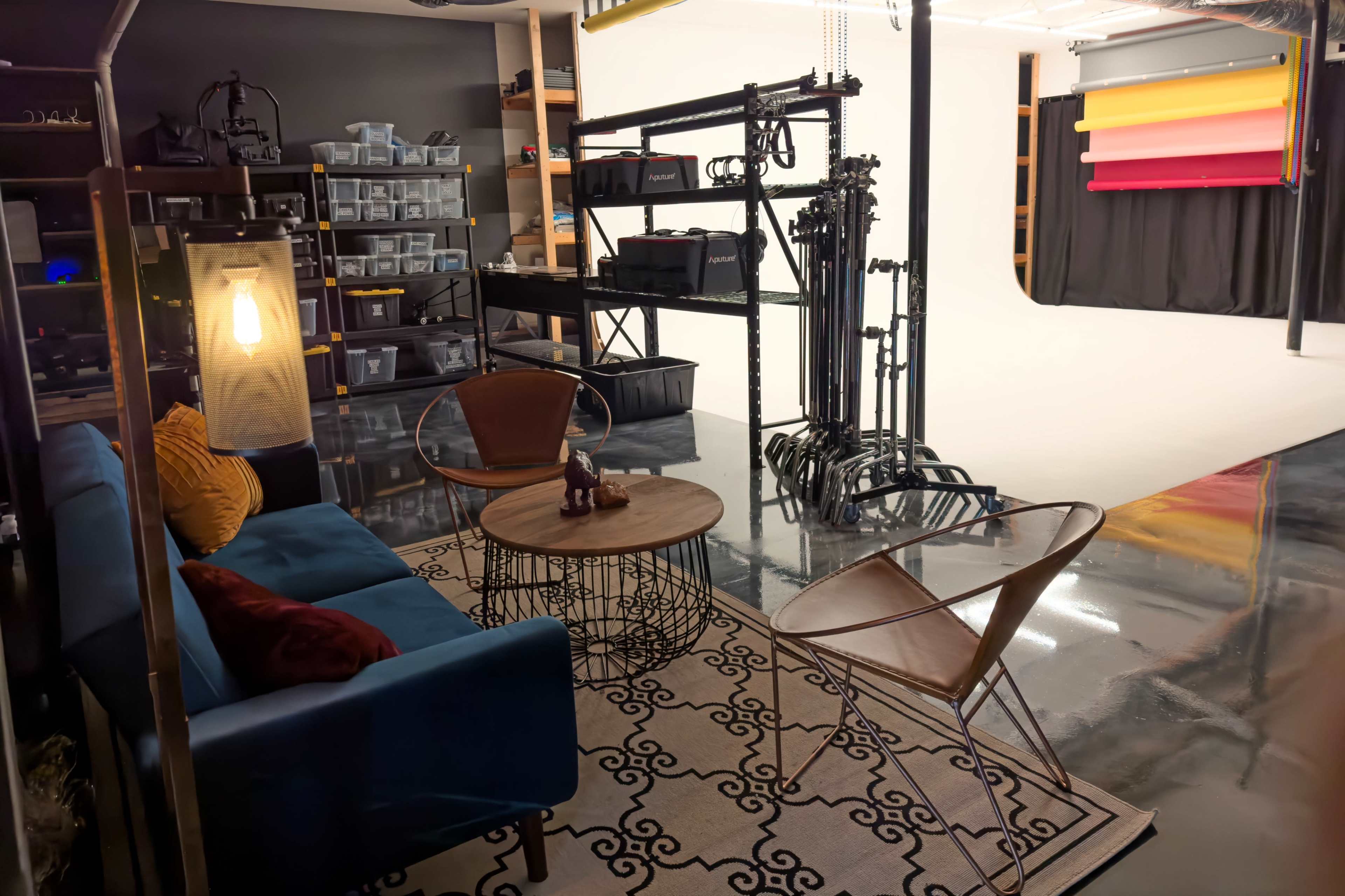 The image shows a photography studio setup with a cozy seating area featuring a blue couch and a round coffee table, alongside various studio equipment and a backdrop in the background.