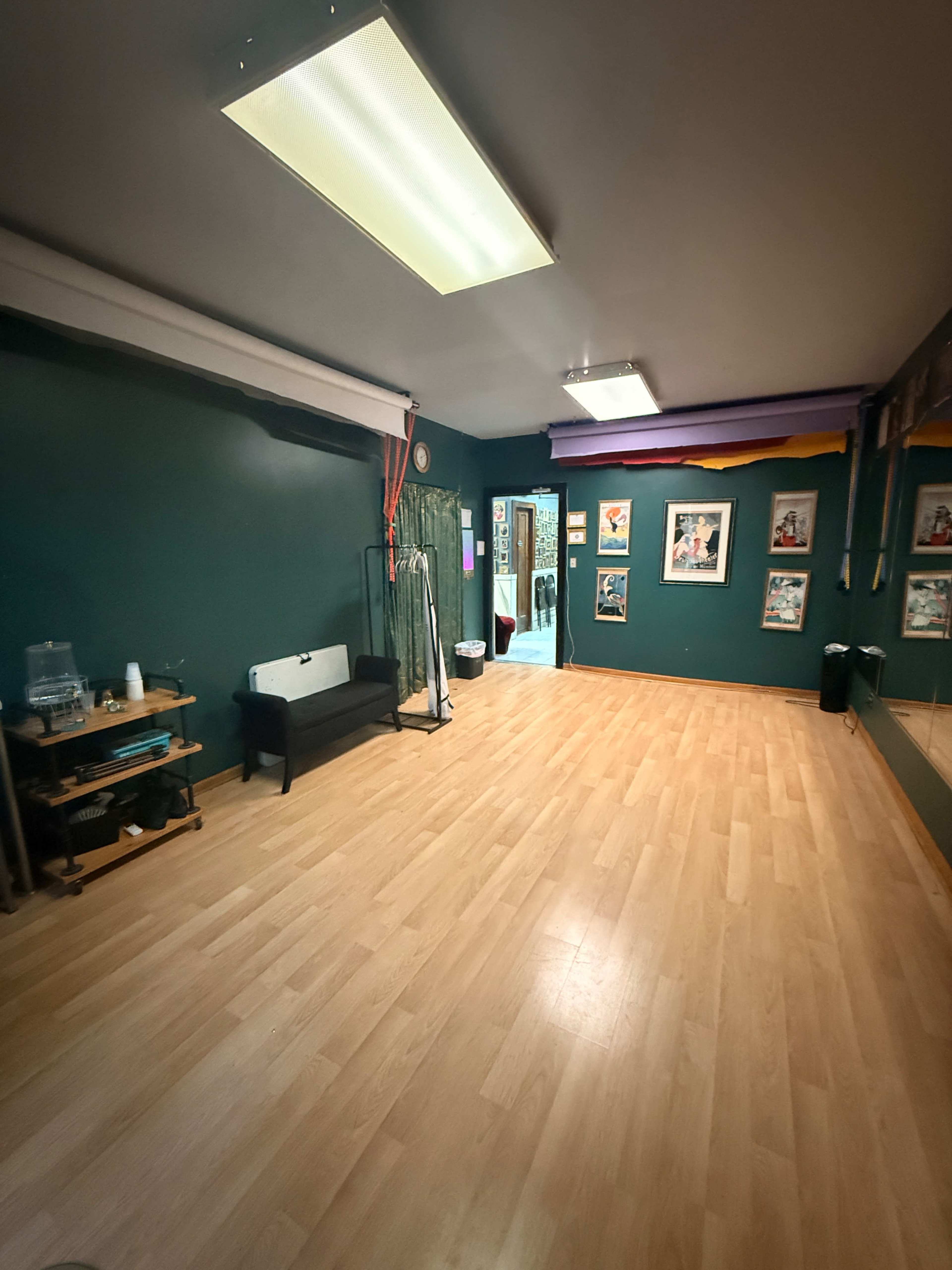 The image shows an empty studio space with wooden flooring, green walls, and various framed pictures hanging on the far wall.