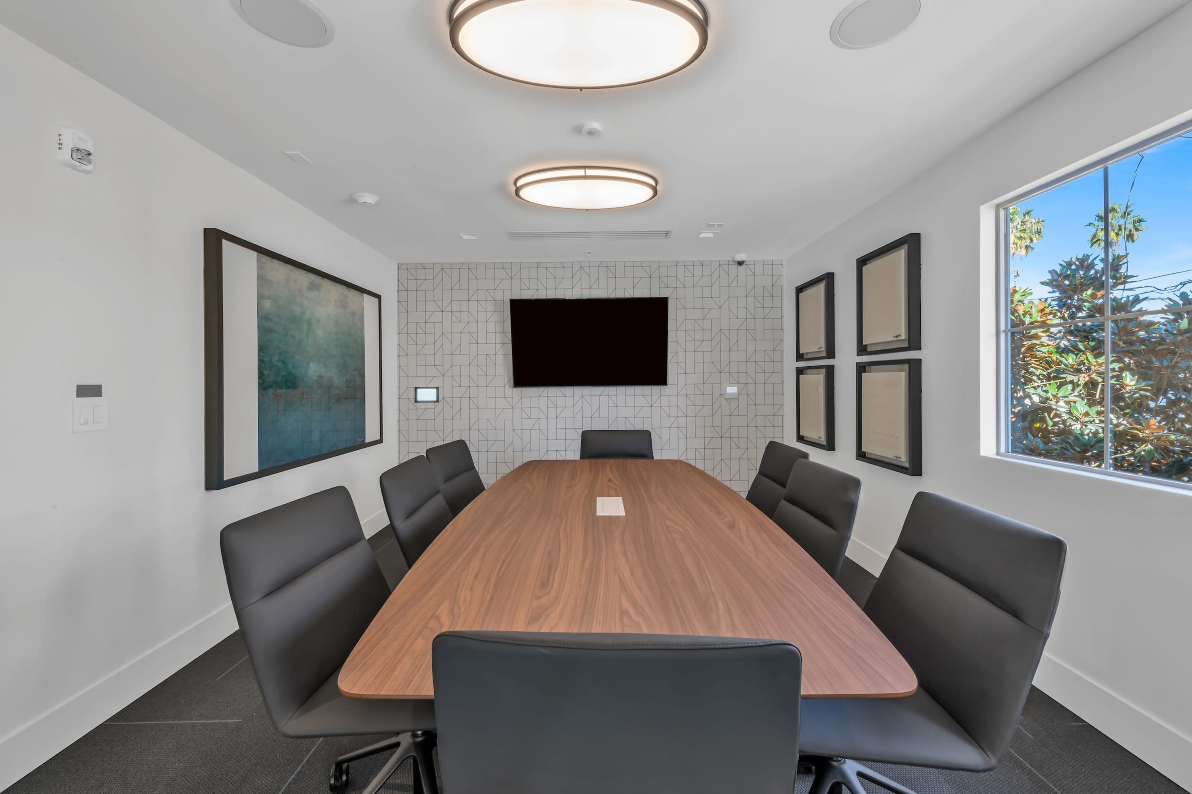 A modern conference room features a long wooden table surrounded by black chairs, with a large screen on the wall and ample natural light from a window.