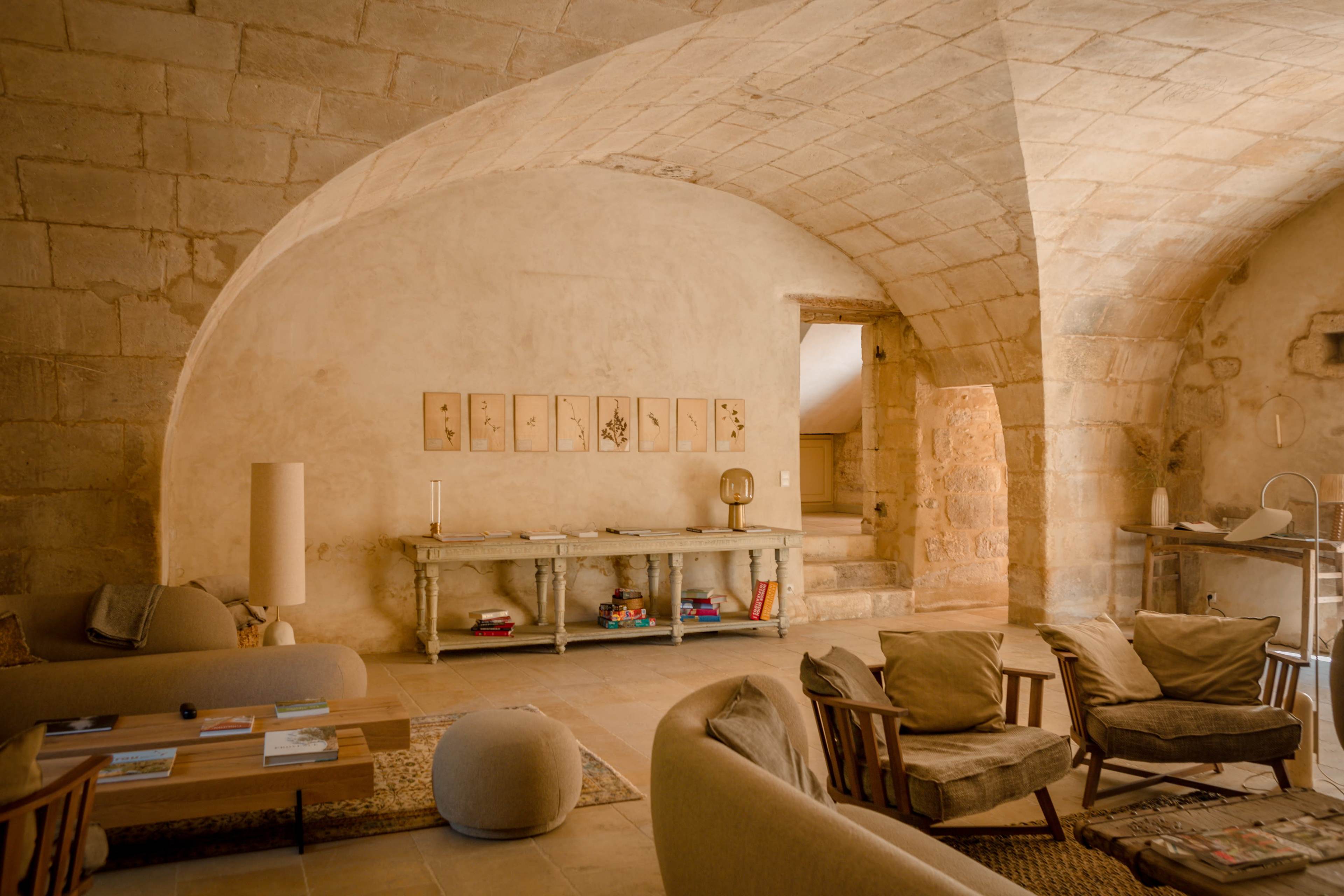 The image depicts a spacious, warmly lit living room with stone walls, arched ceilings, and a mix of modern and rustic furniture arranged around a central coffee table.