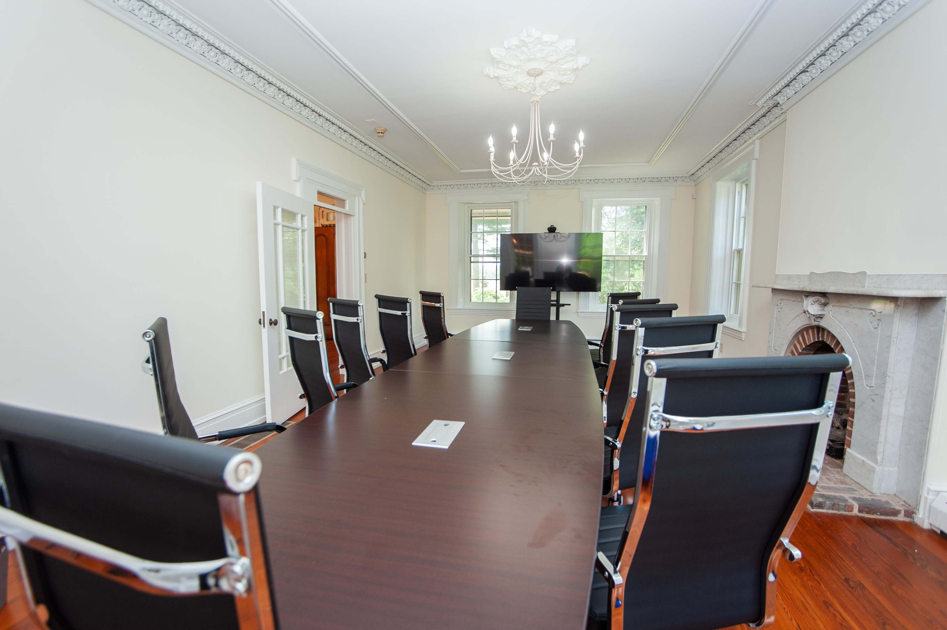 The image shows a large, formal conference room with a long wooden table, black chairs, a television screen, and a chandelier.