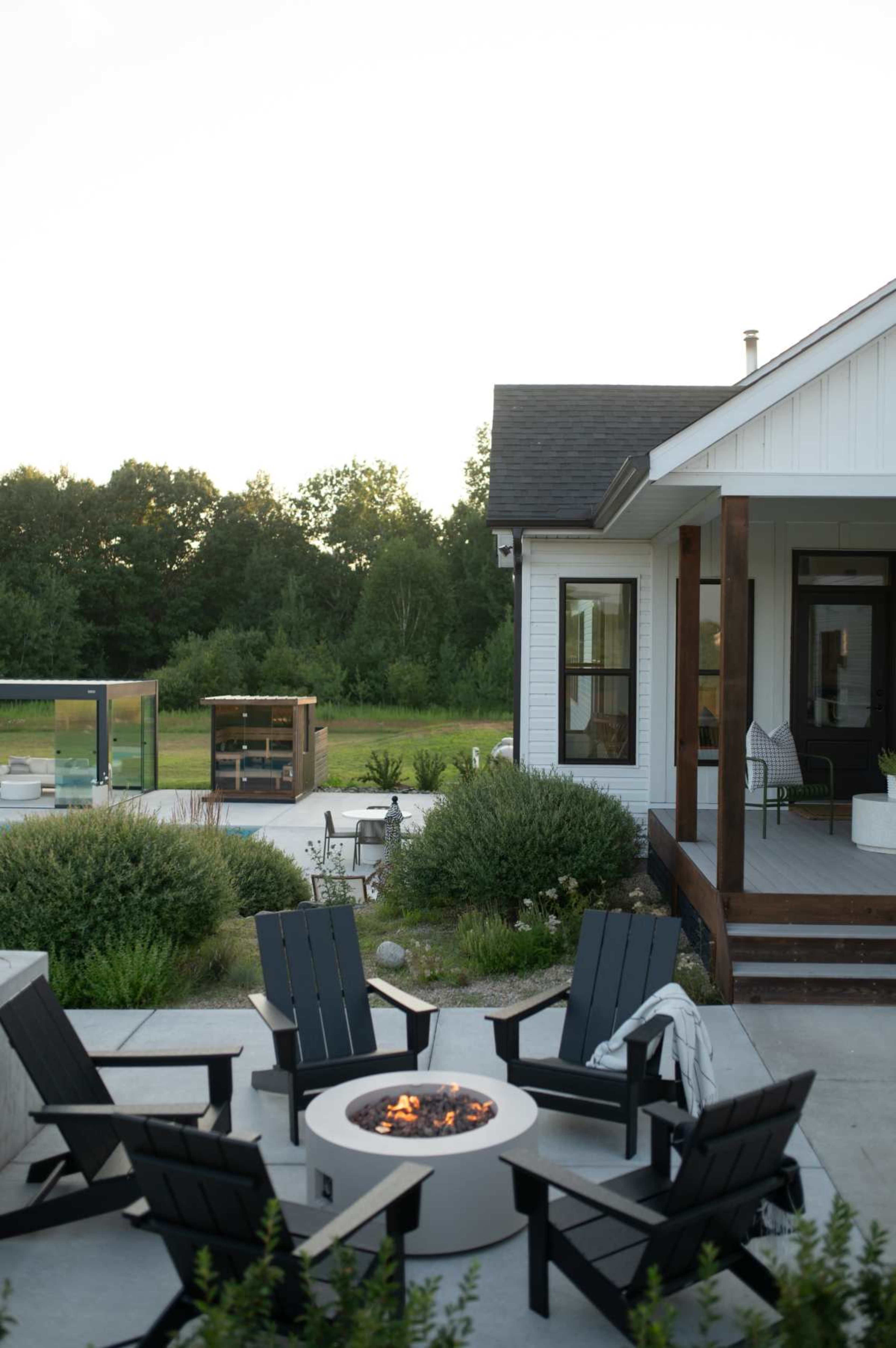 A modern patio features a circular fire pit surrounded by black Adirondack chairs, next to a grassy lawn and a house with a porch.