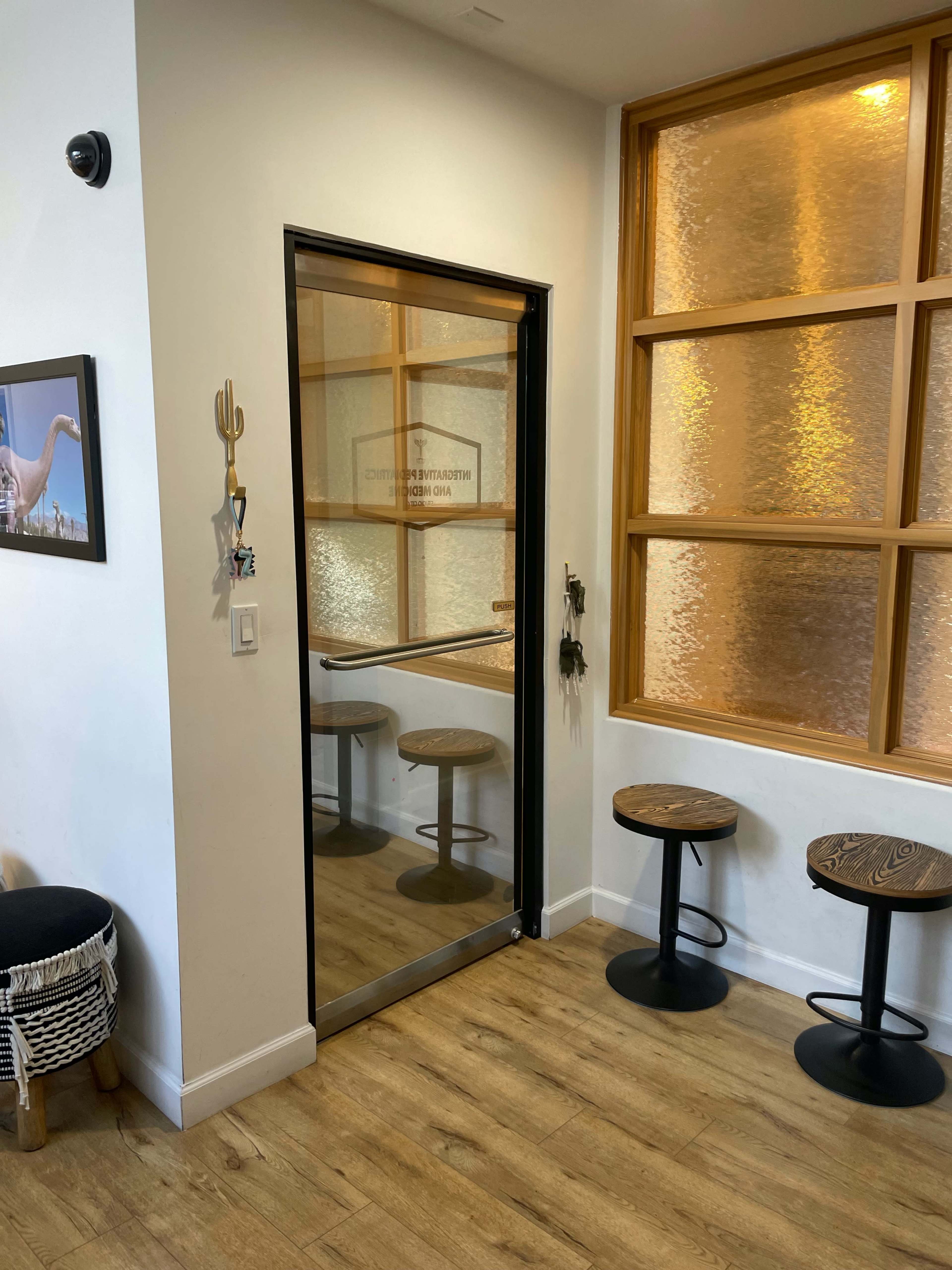A small interior corner featuring a glass door, a wall with a framed picture, and two round stools beside a large window with frosted glass.