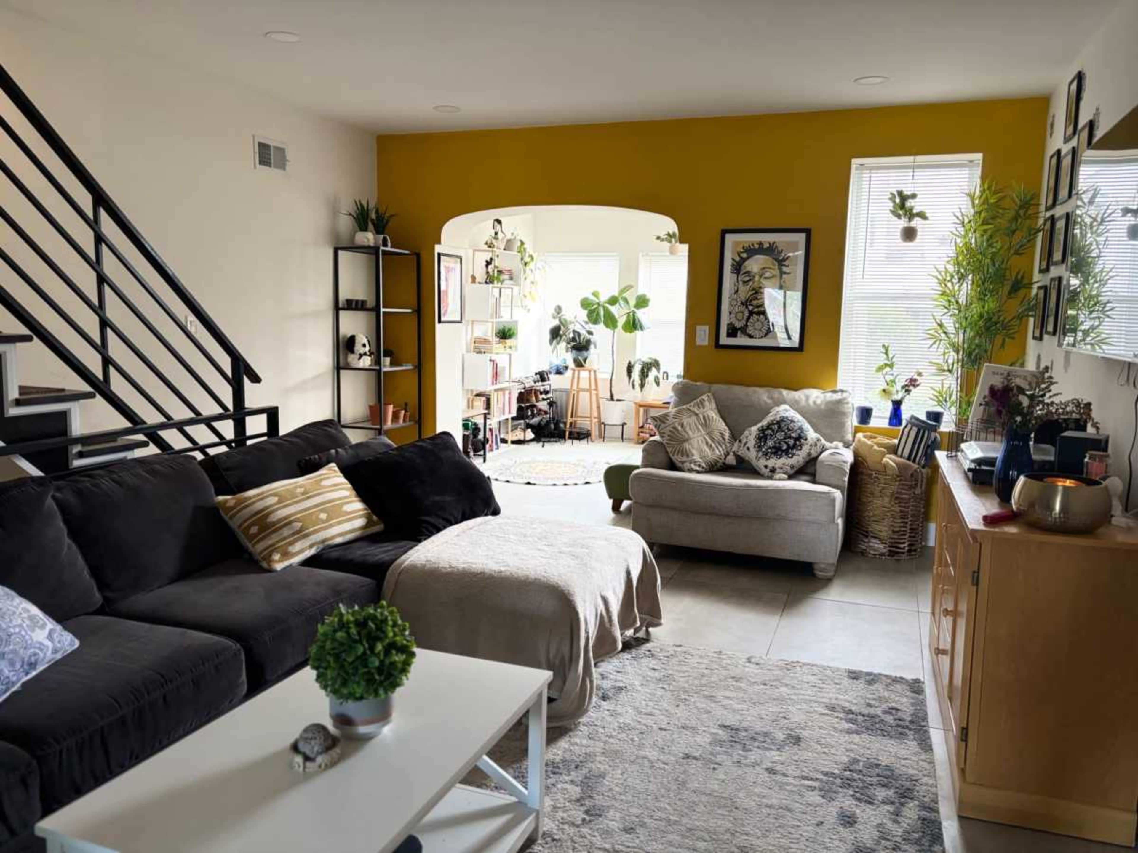 The image shows a bright and open living area featuring a dark sofa, a light-colored armchair, and a wooden media console, with plants and artwork adorning the walls.
