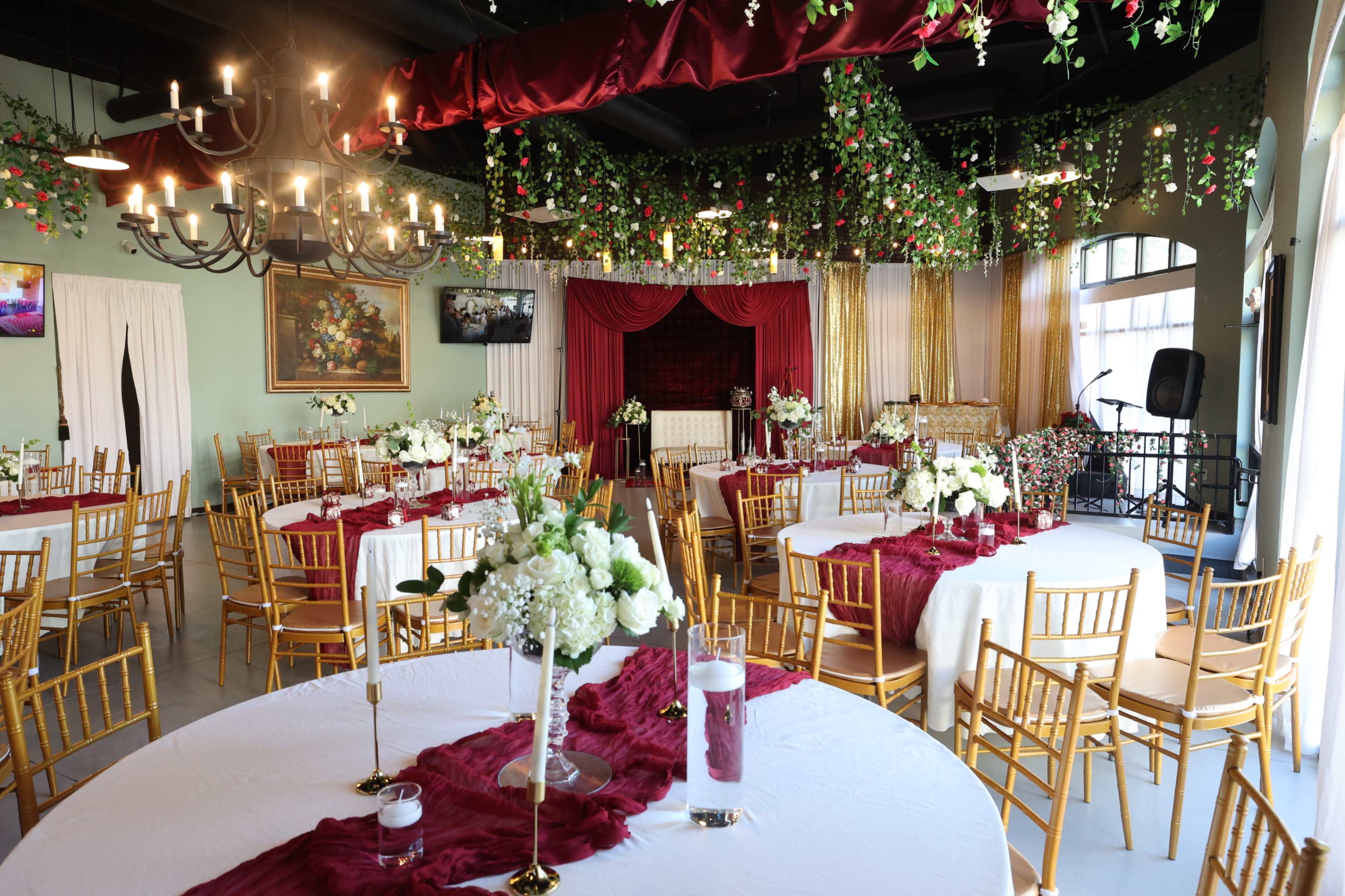 The scene depicts an elegantly decorated banquet hall with round tables covered in white tablecloths, gold chairs, and lush greenery hanging from the ceiling.