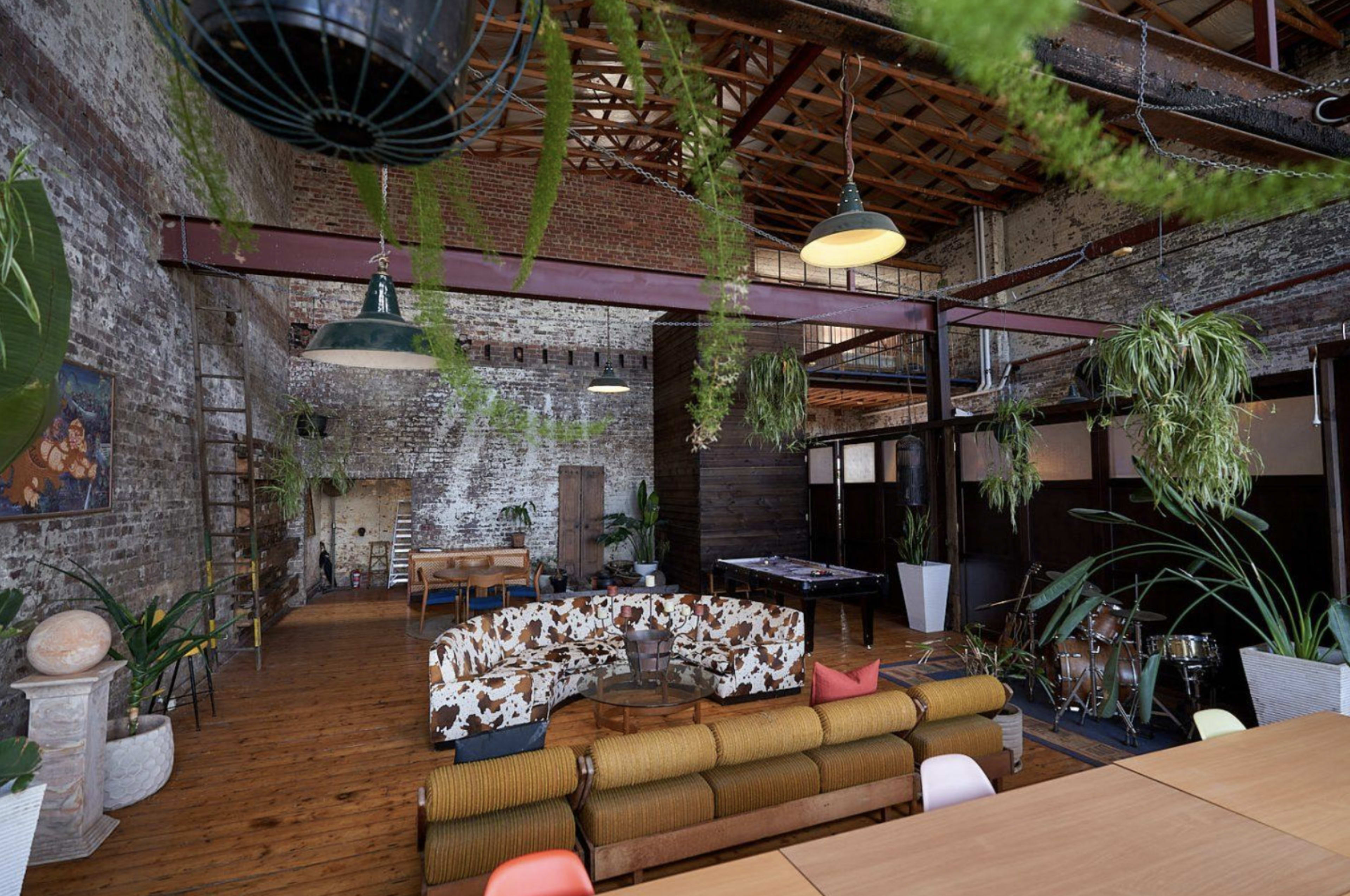 A spacious interior featuring exposed brick walls, wooden flooring, and modern furnishings, including a large couch and potted plants.