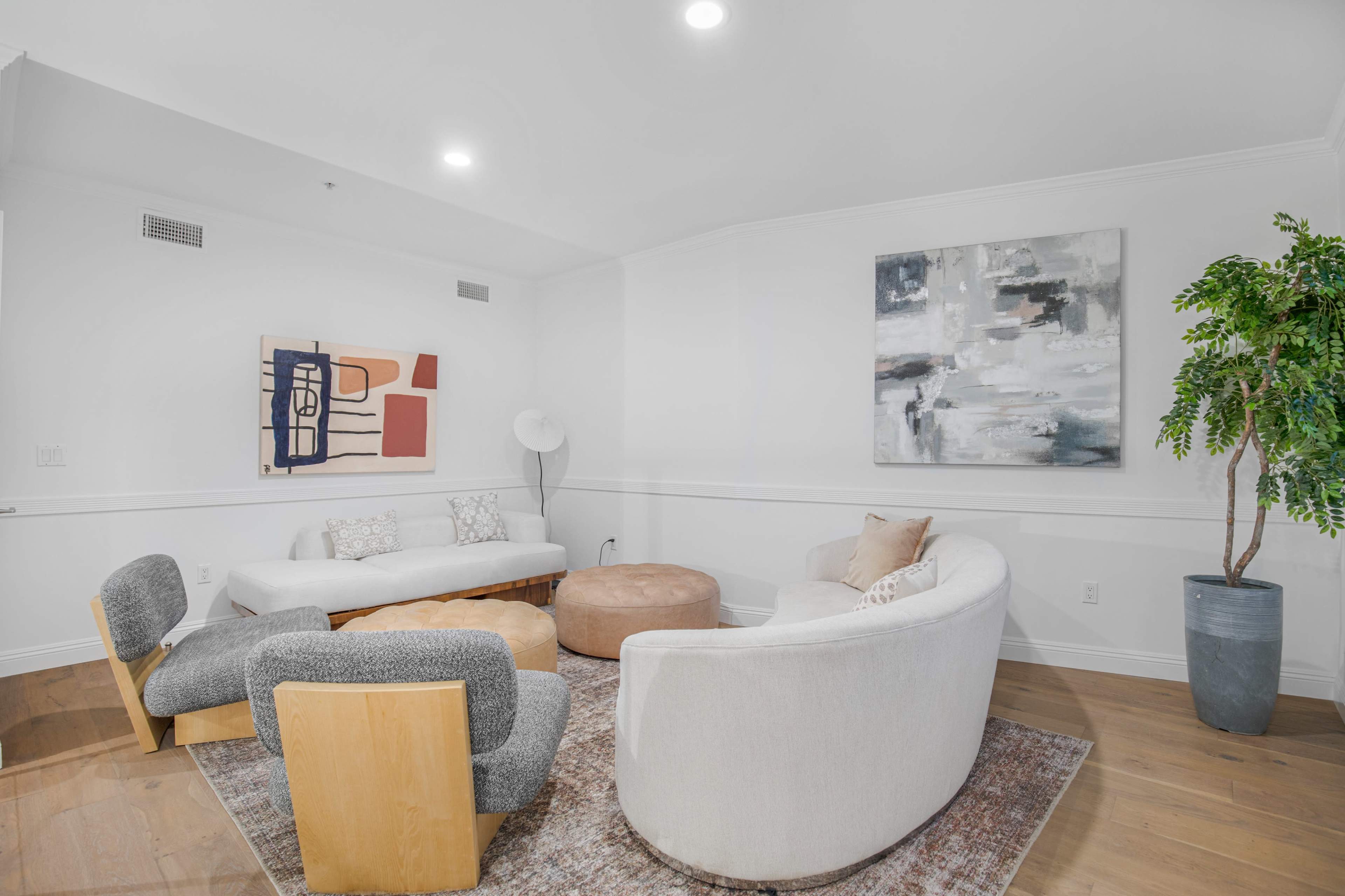 The image shows a modern living room featuring a circular arrangement of upholstered furniture, a large abstract painting, and a potted plant in the corner.