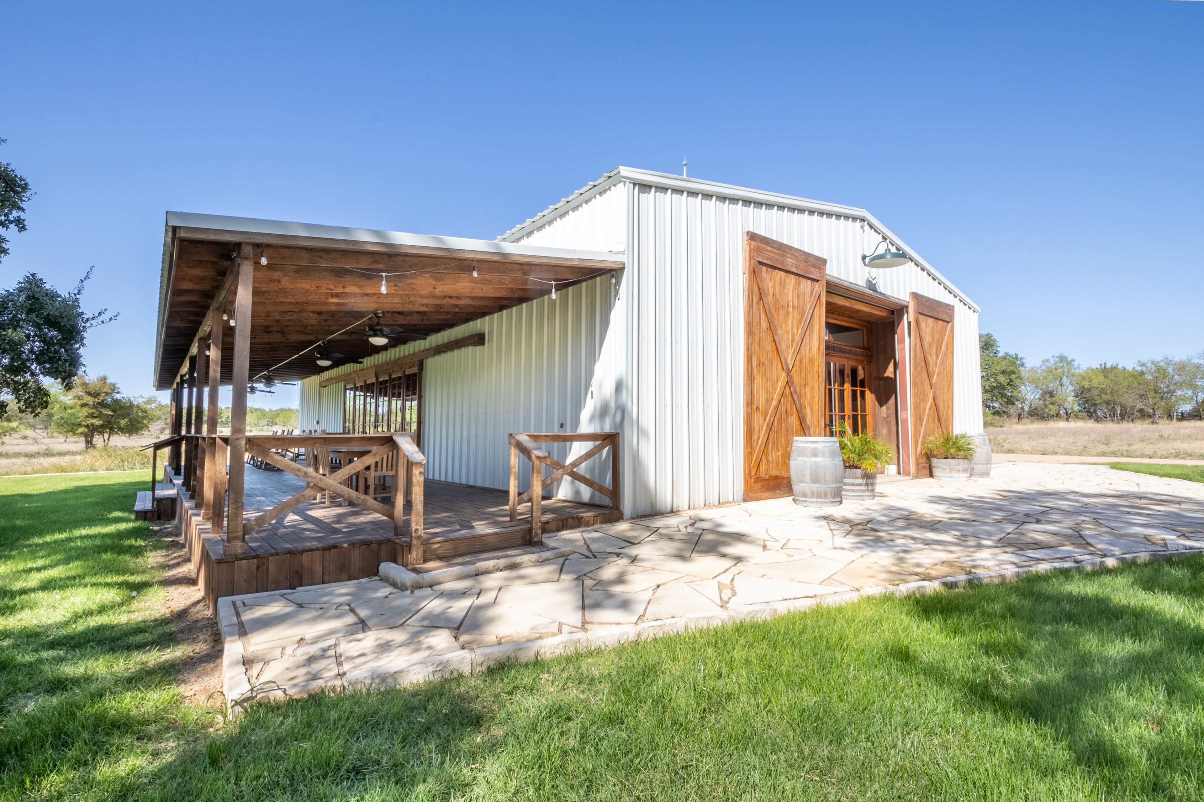 A metal barn with wooden doors and a covered porch, set on a grassy area with stone paving.