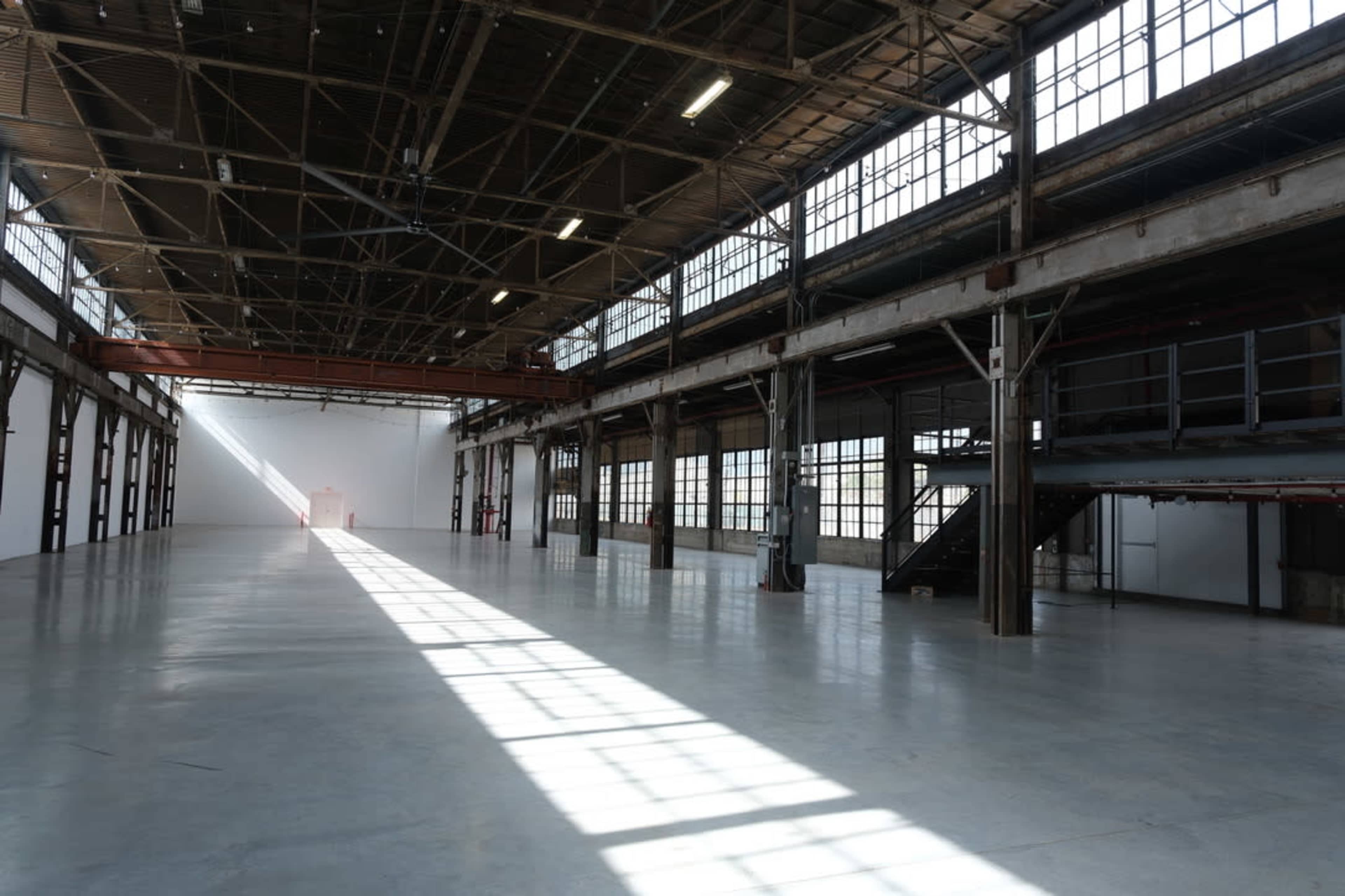 The image shows a large, empty industrial space with high ceilings, large windows, and concrete flooring.