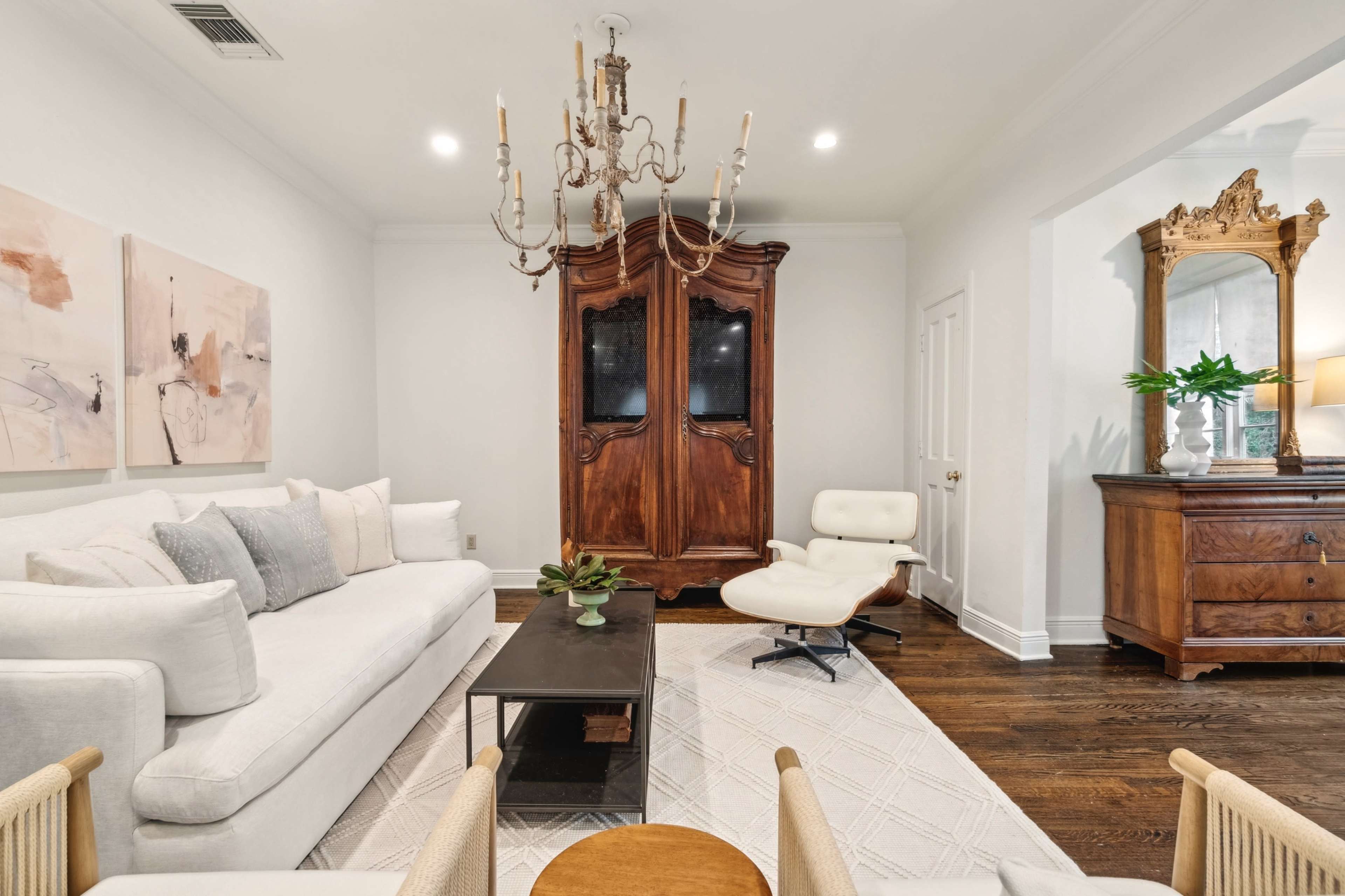 The image shows a stylish living room featuring a white sofa, a wooden armoire, a modern chair, and decorative elements on a coffee table.