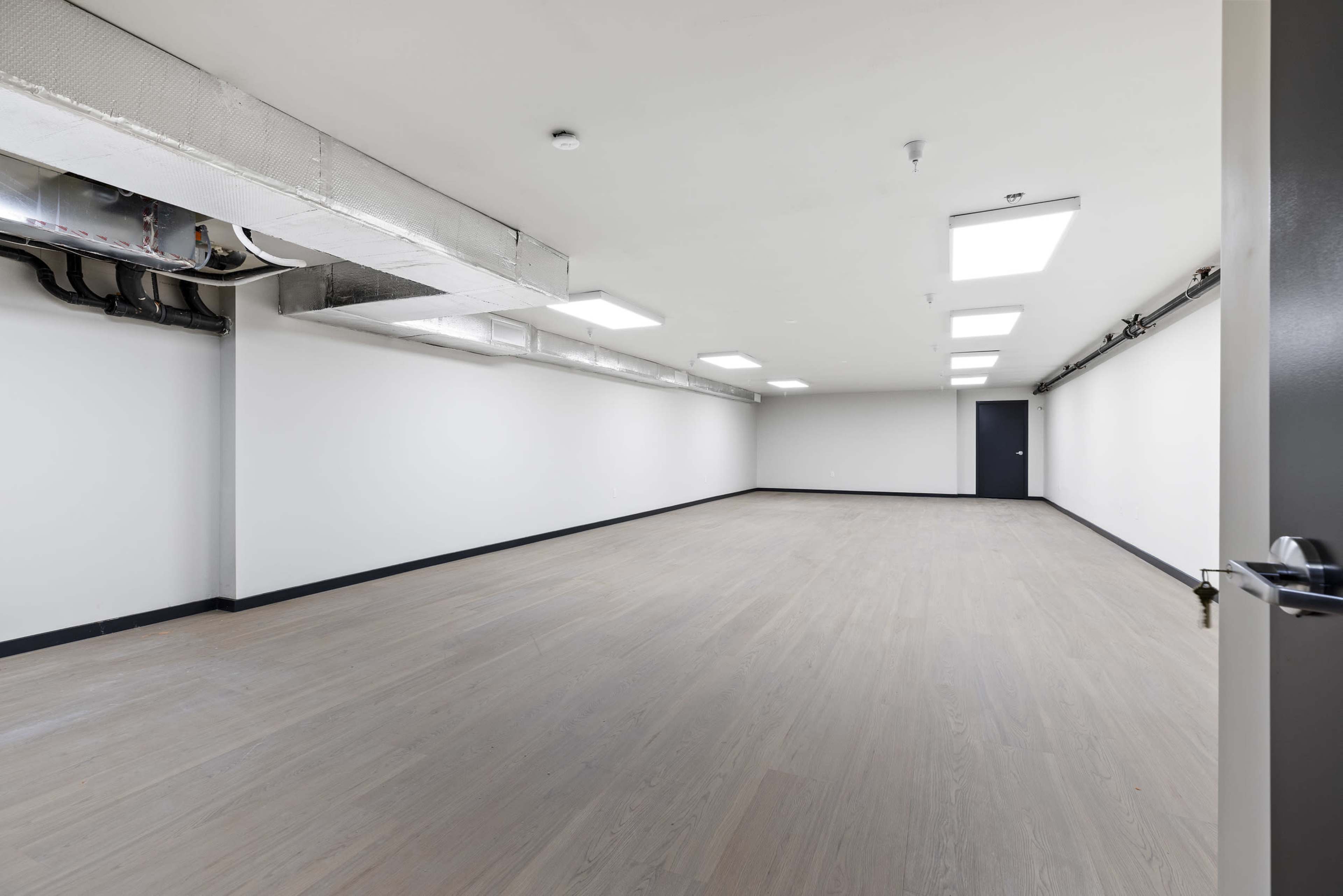 The image shows a large, empty room with white walls, a light-colored floor, and overhead lighting.