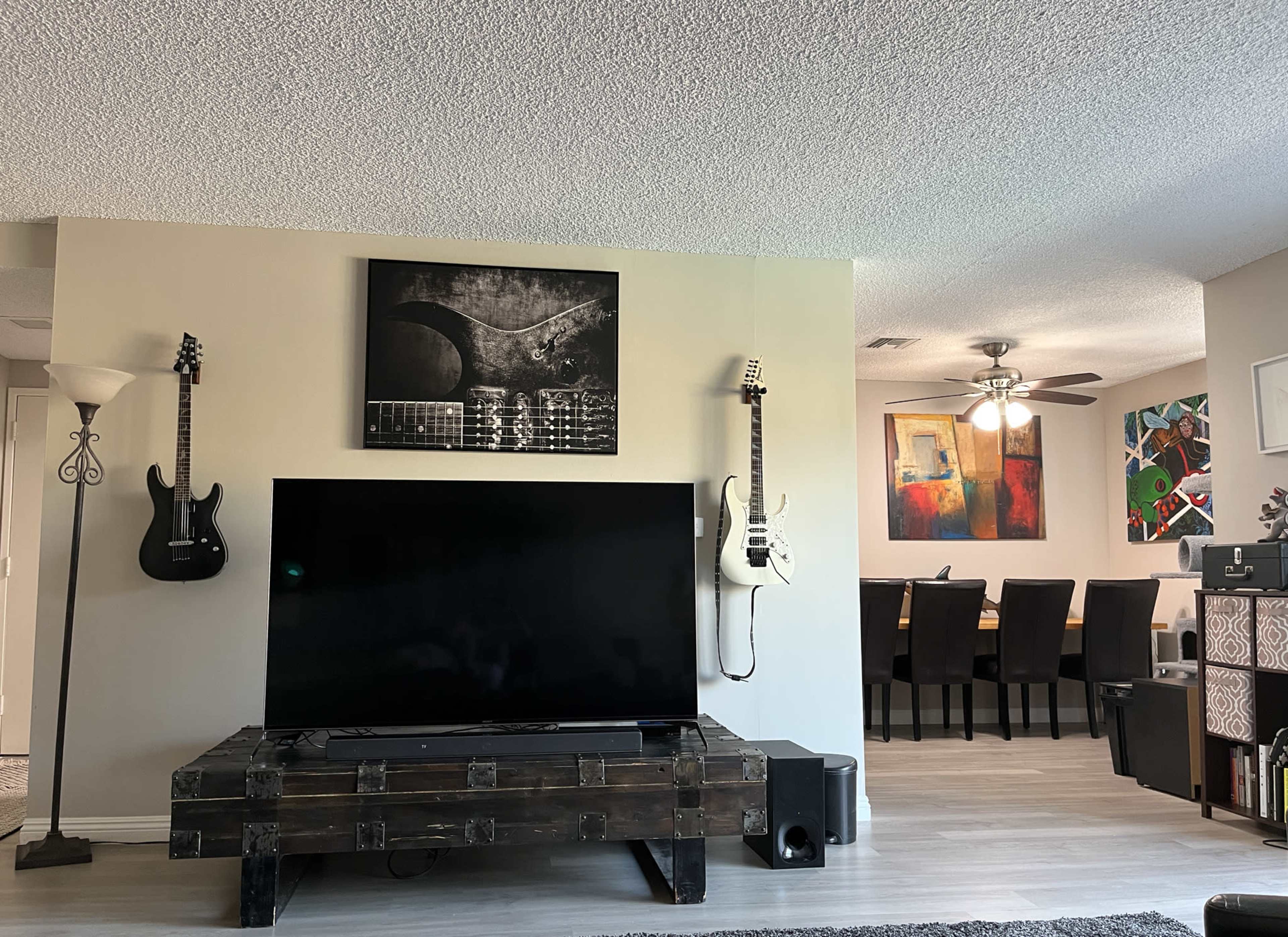 A living room with a large flat-screen TV on a wooden stand, flanked by two electric guitars mounted on the wall, and a dining area visible in the background.