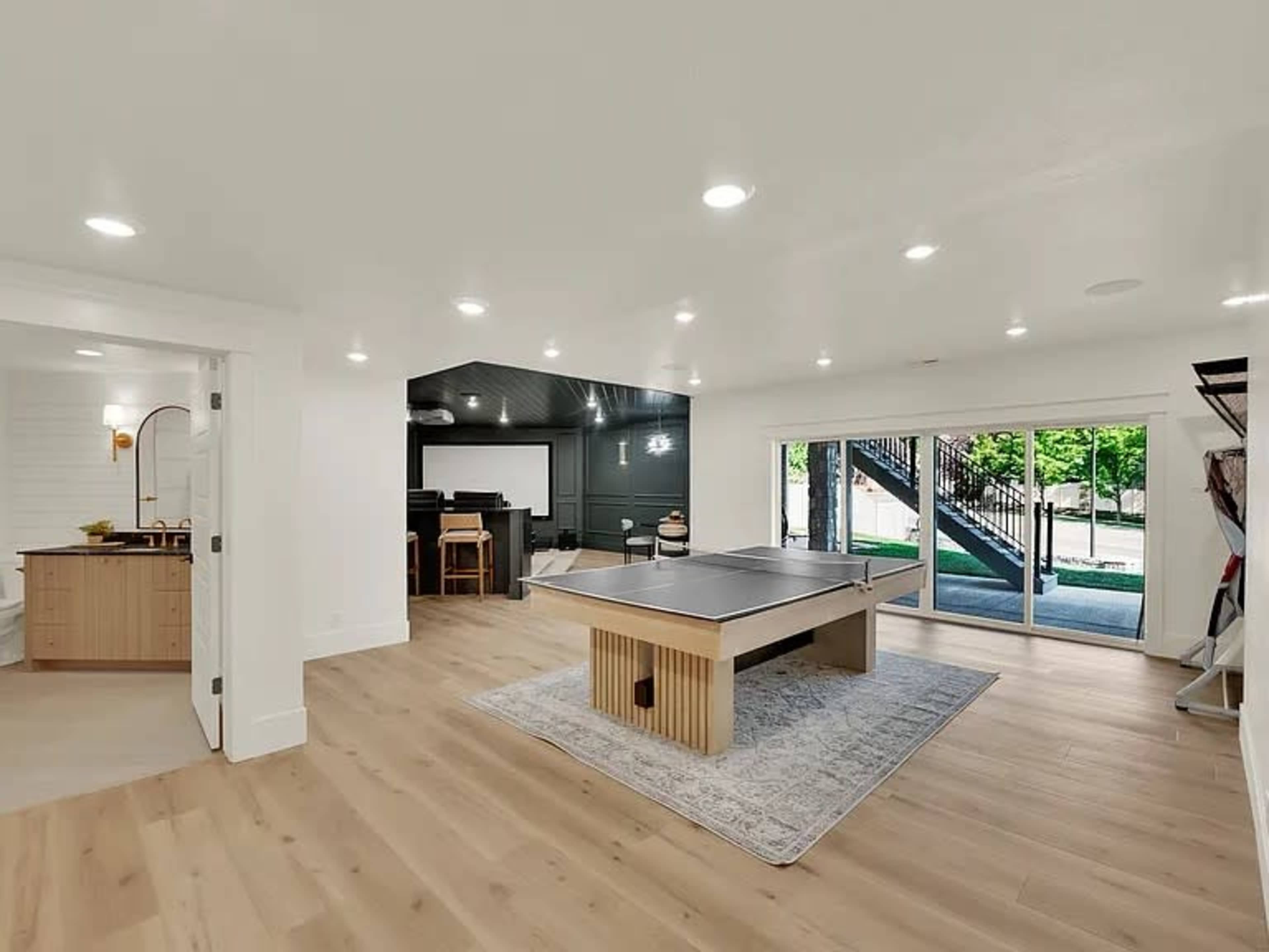 The image shows a modern basement interior featuring a pool table on a rug, with sliding glass doors leading outside, and a kitchenette area visible to the left.