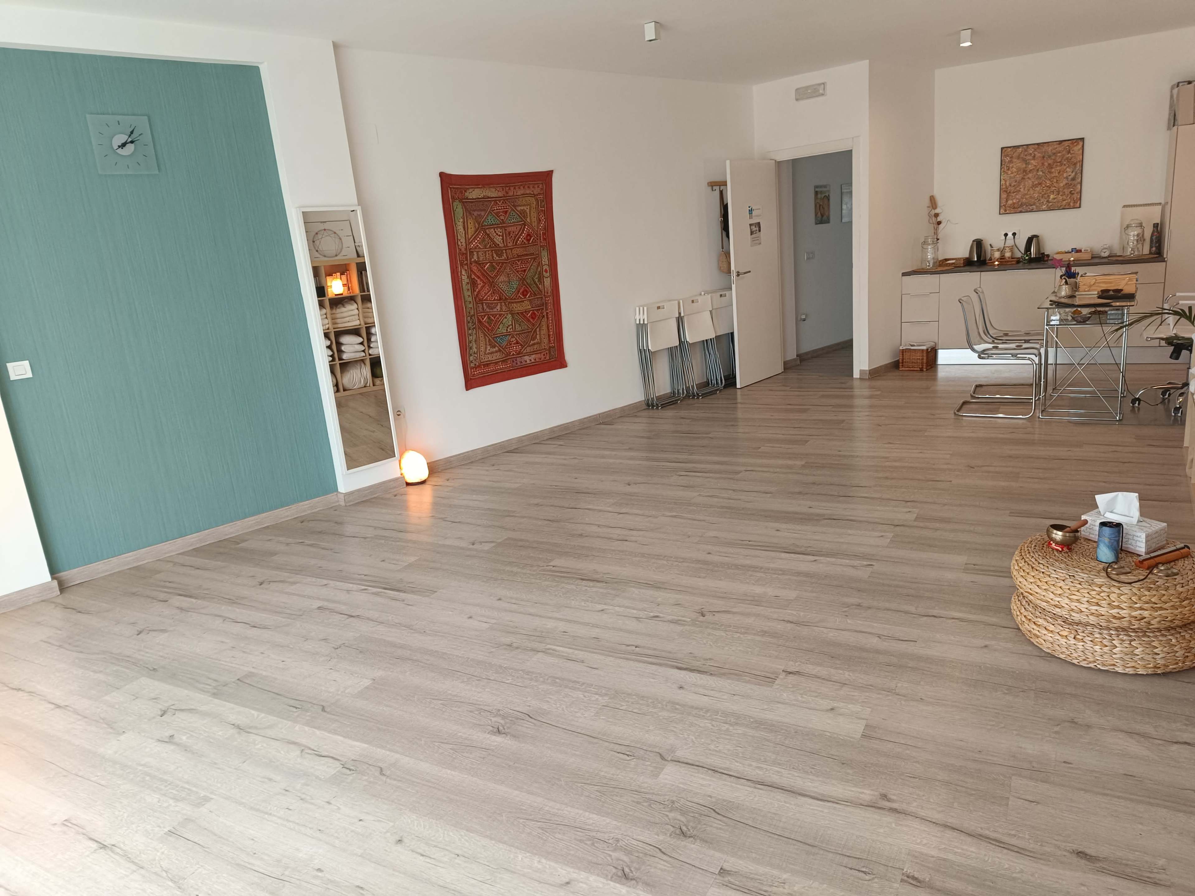 The image shows a spacious, vacant room with wooden flooring, minimal furniture, and a decorative wall hanging.