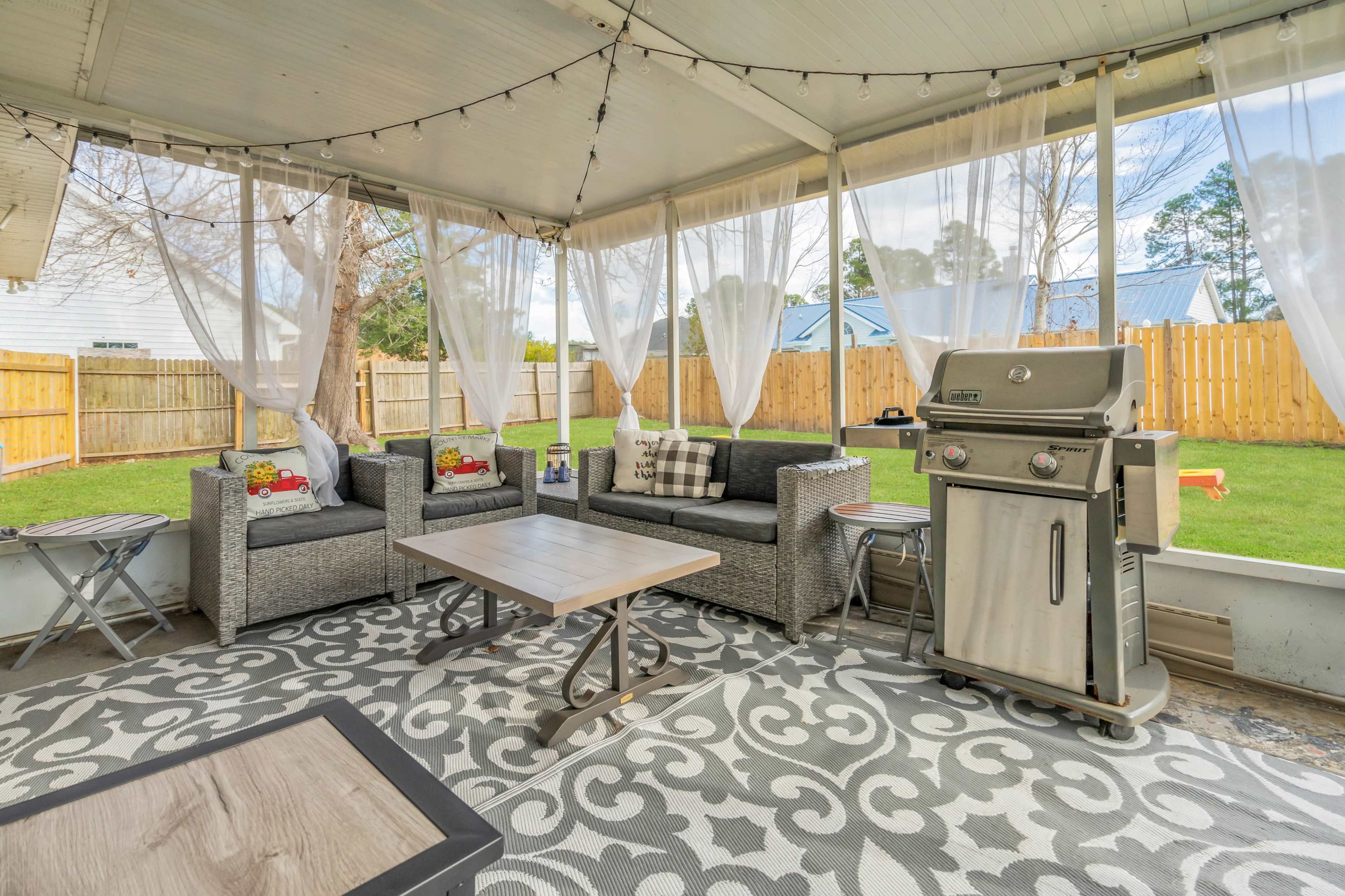 A covered outdoor patio featuring a grill, seating area with gray couches, a coffee table, and a decorative rug, all surrounded by a backyard with a wooden fence.