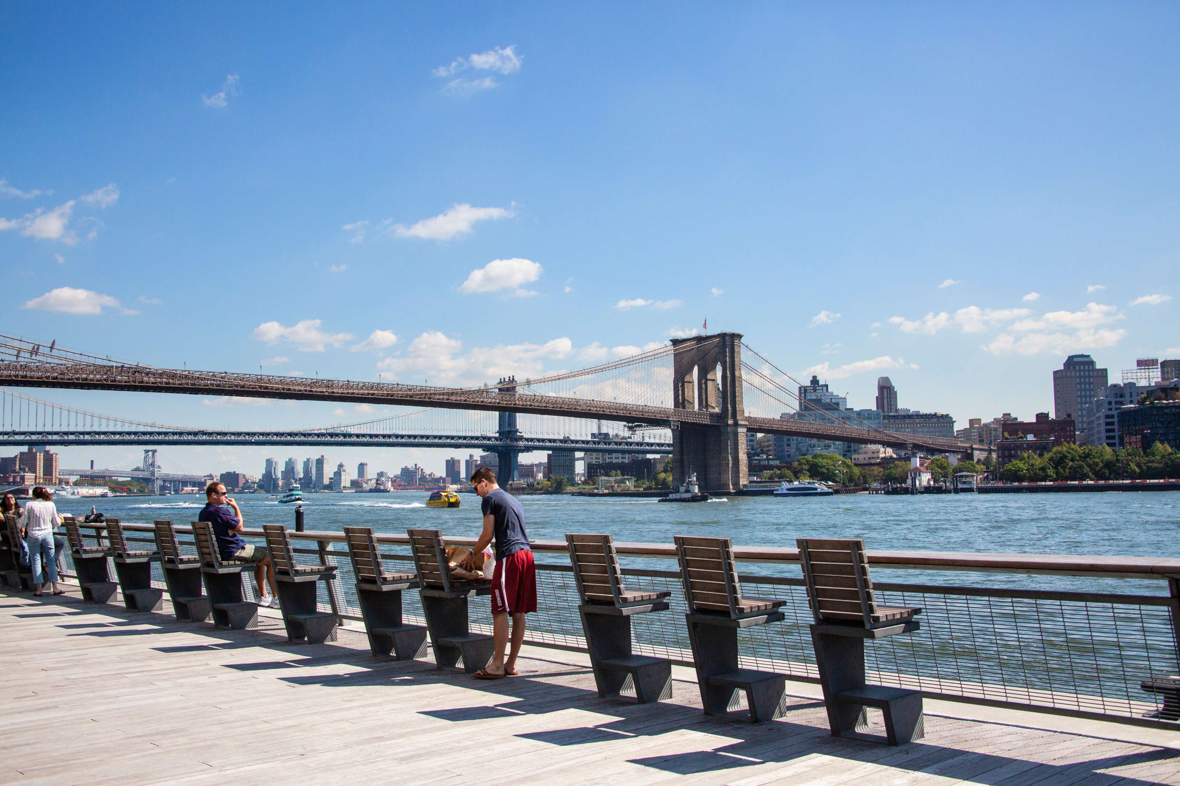 A waterfront scene in New York City with people sitting on a deck, facing the Brooklyn Bridge and the East River.