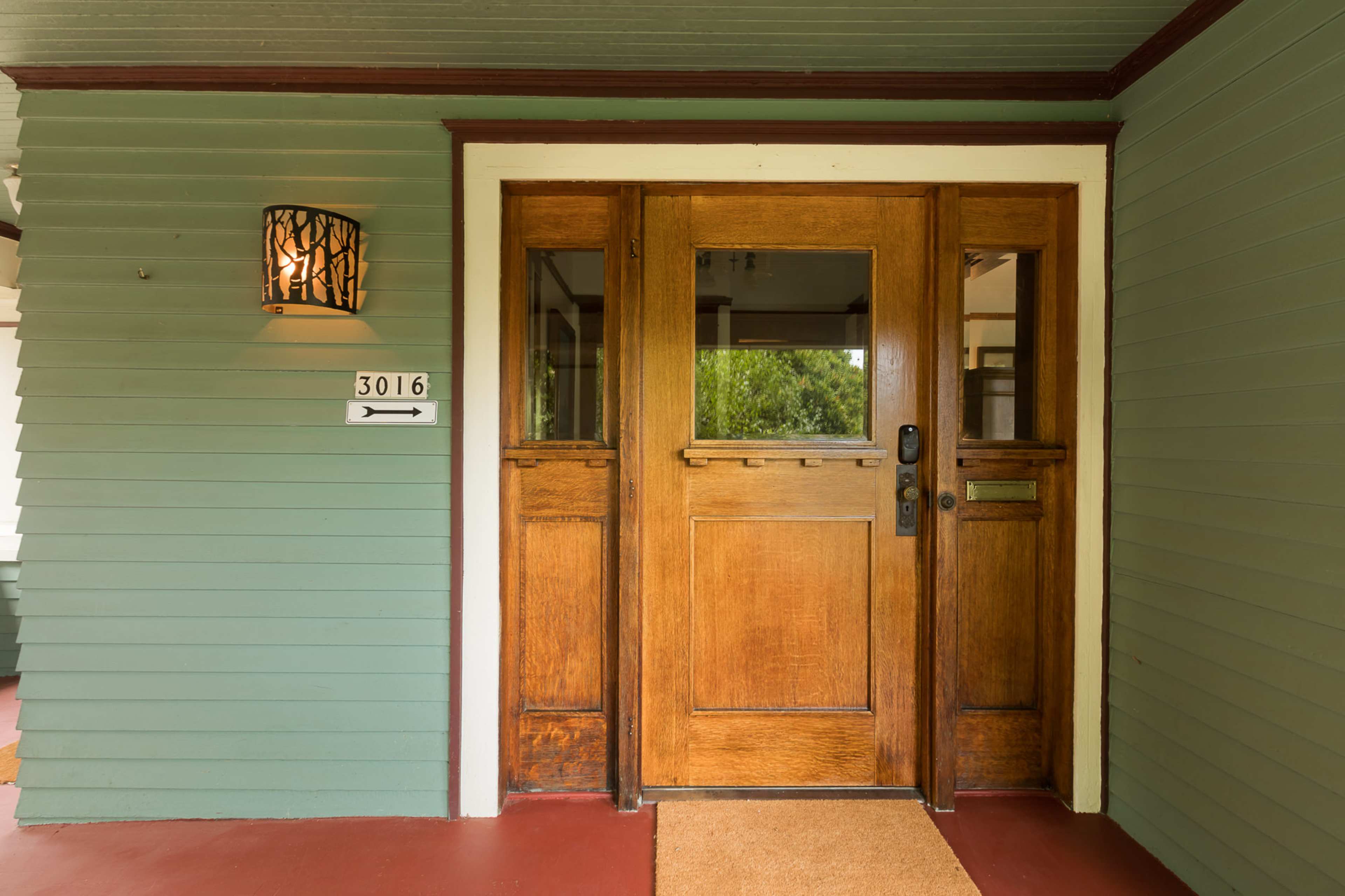 The entrance features double wooden doors with glass panels, flanked by a wall-mounted light fixture and a house number mounted to the side.