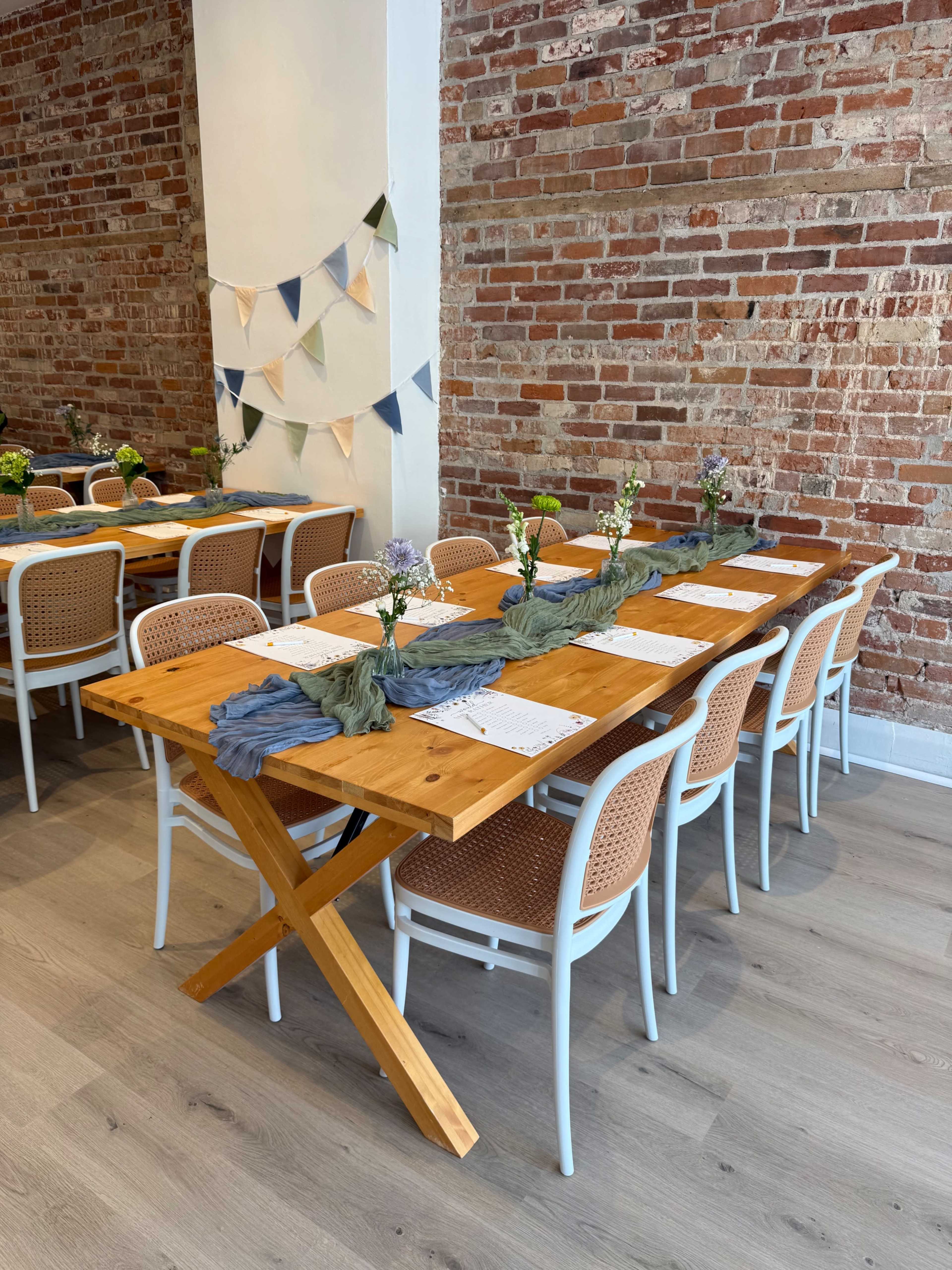 A long wooden table is set with decorative elements in a modern dining space featuring exposed brick walls and light-colored chairs.