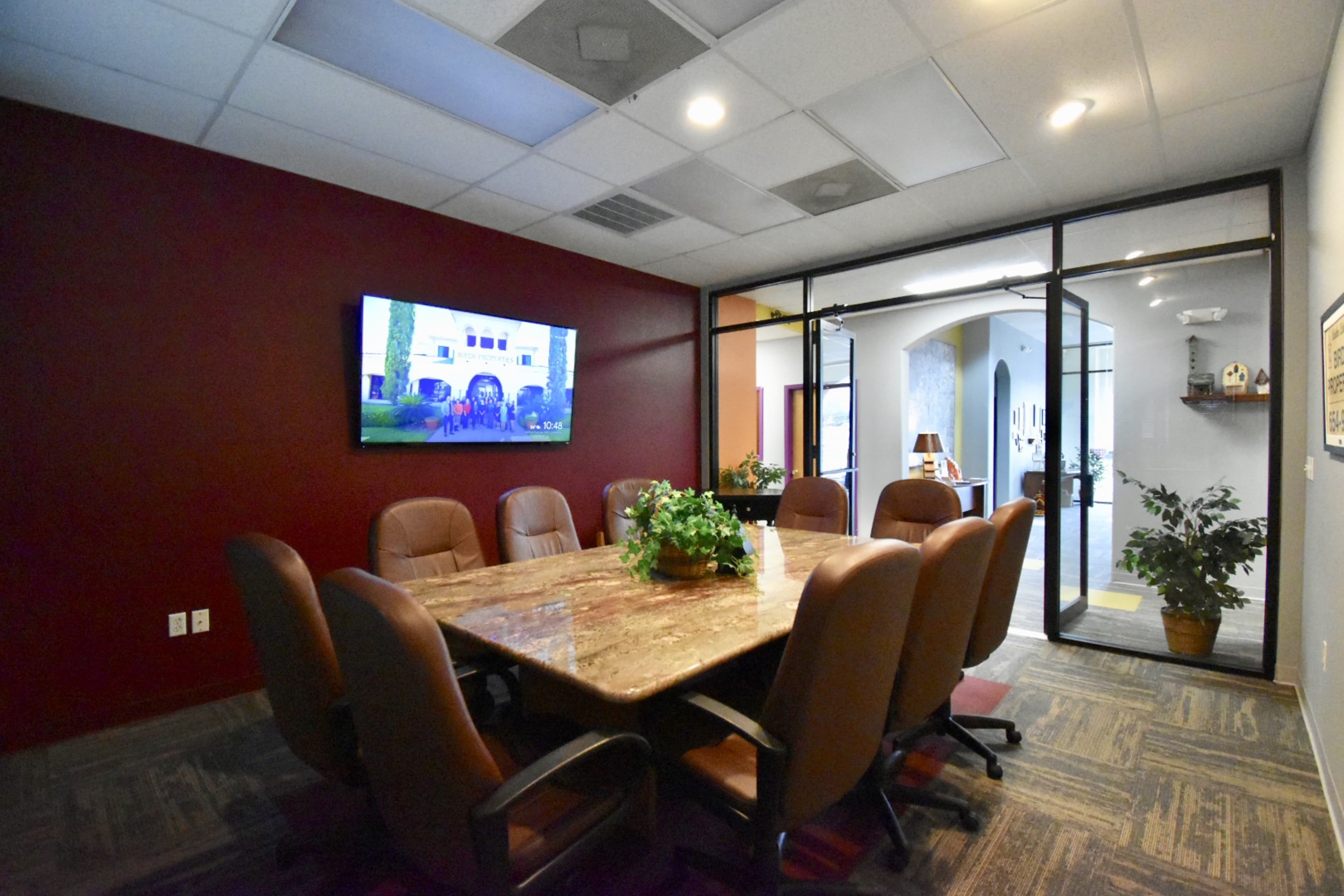 A conference room features a large table surrounded by leather chairs and a wall-mounted TV in a space with dark red walls.