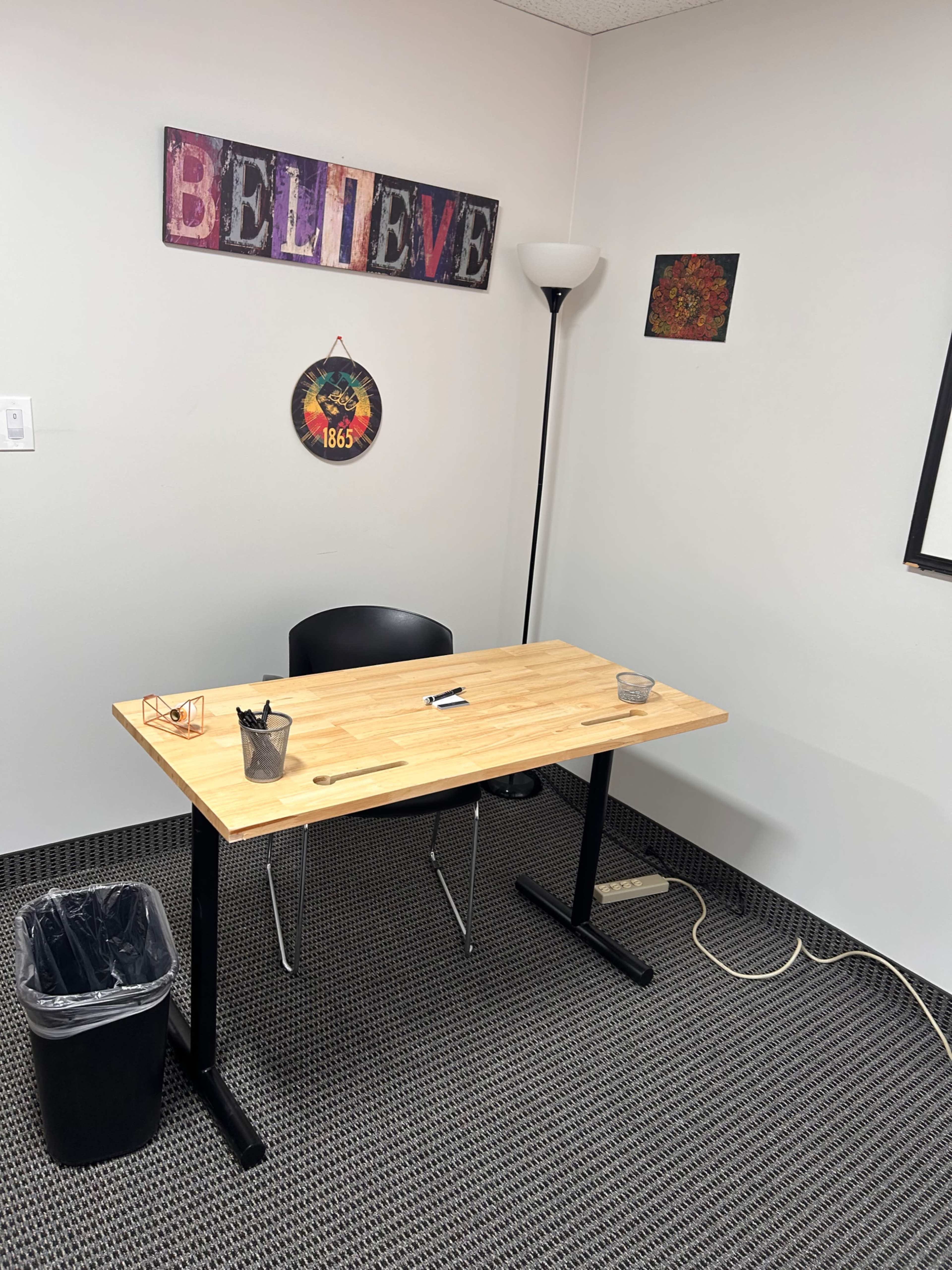 A wooden desk with a chair, glasses, a pen, and two containers sits in an empty office space with a lamp and wall decorations.