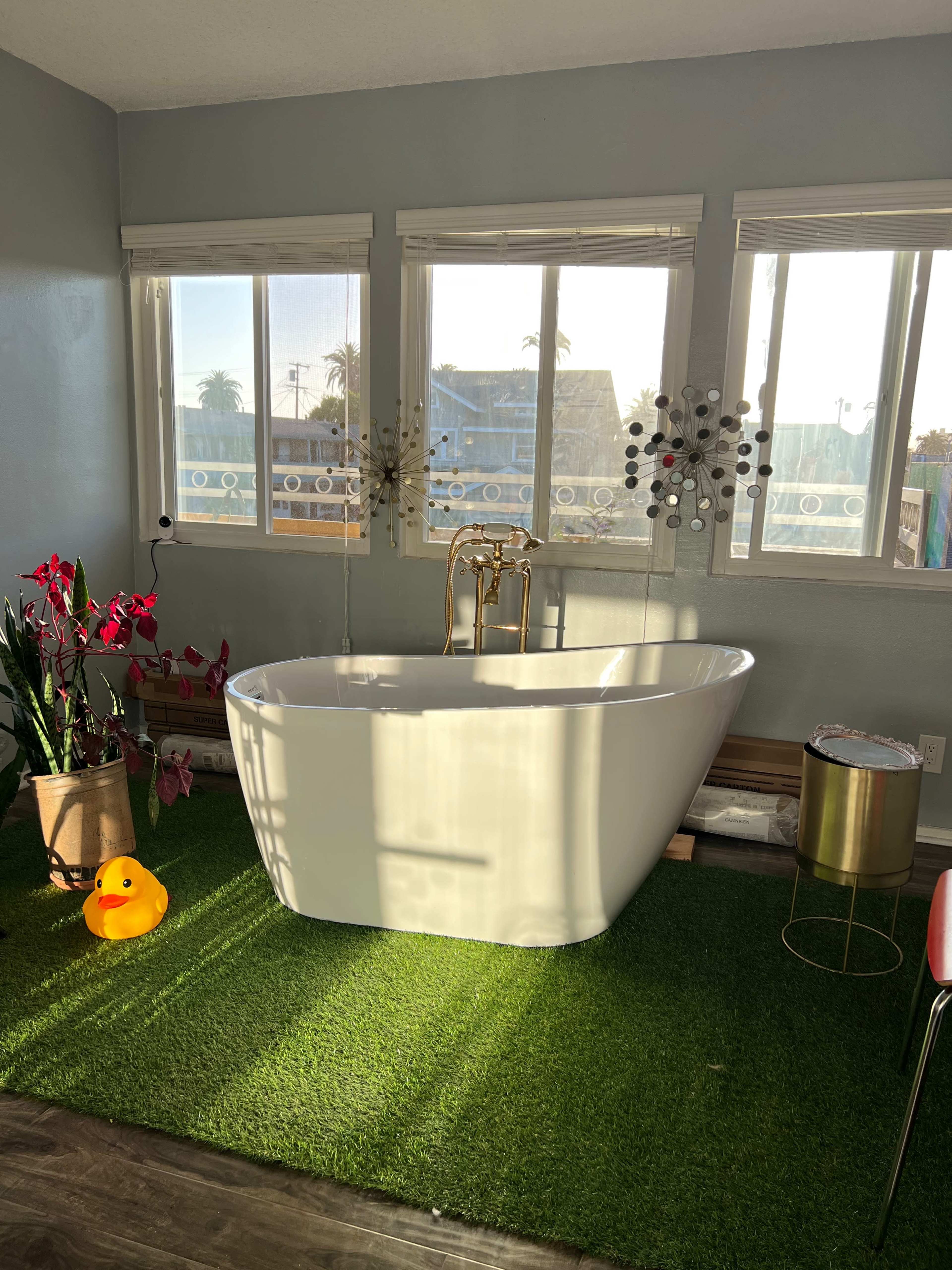A white freestanding bathtub on artificial grass, with a rubber duck, a gold-colored side table, potted plants, and large windows allowing natural light to illuminate the space.