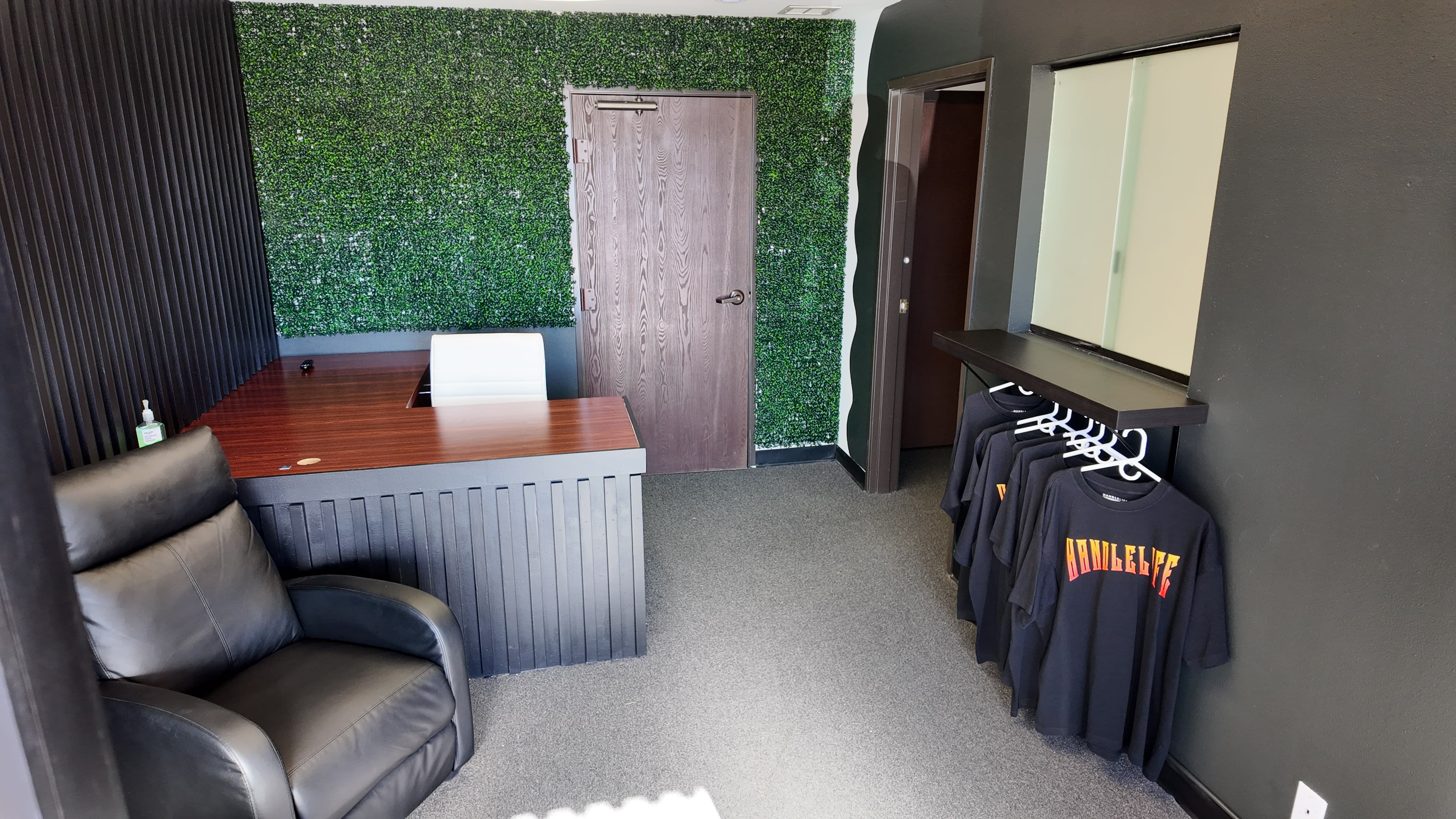 The image shows a small office space featuring a desk, a black leather chair, a green wall with faux foliage, and a clothing rack displaying black T-shirts.
