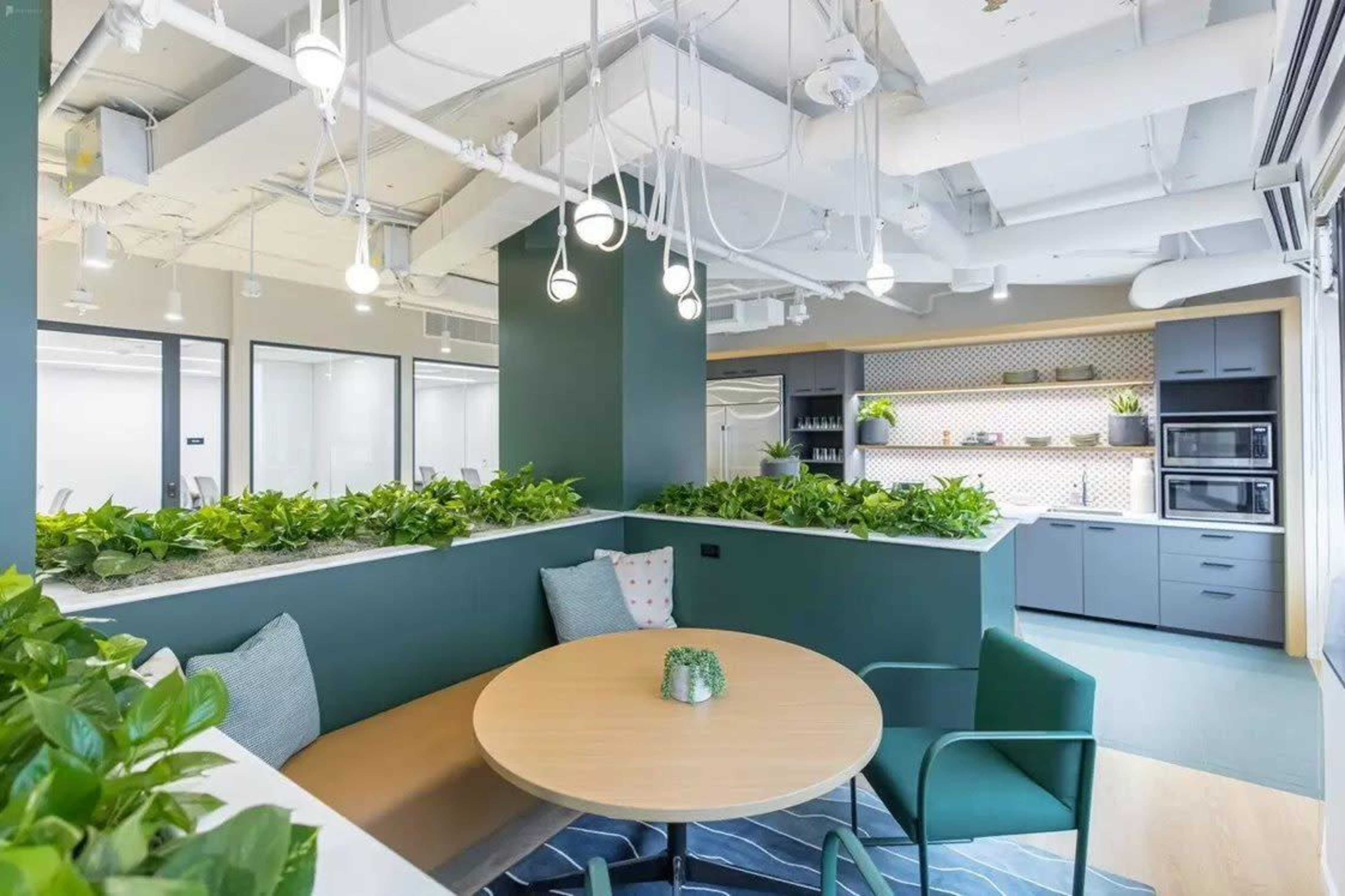 A modern office space features a seating area with a round table surrounded by green plants, adjacent to a kitchen with dark cabinetry and appliances.